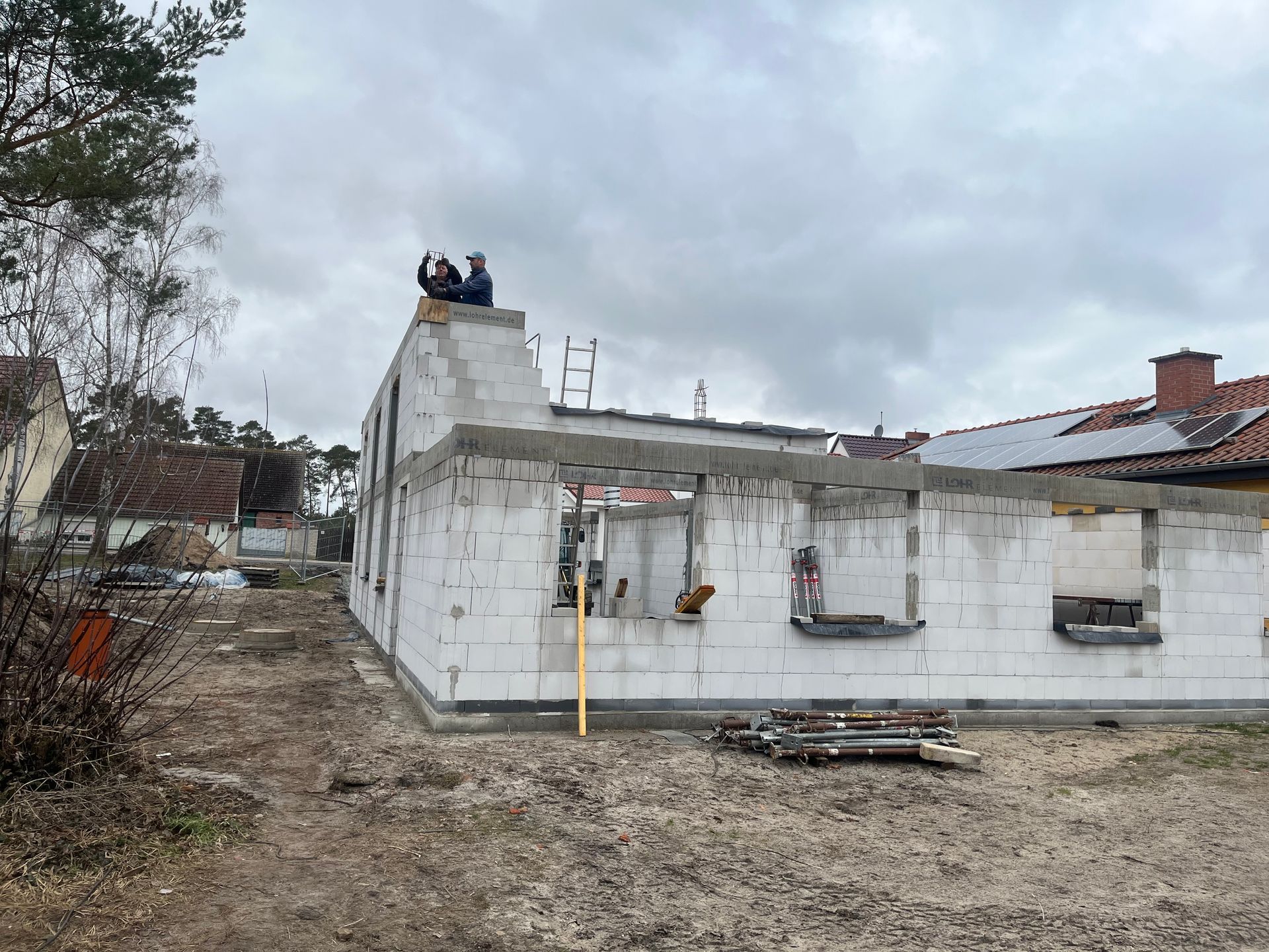Baustelle mit Mauern aus Zementblöcken, zwei Personen auf dem Dach. Bewölkter Tag.