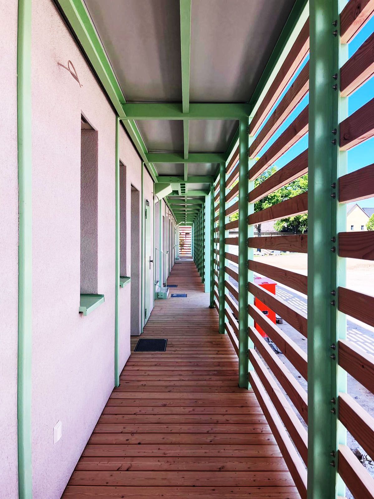 Holzsteg zwischen Gebäuden mit grünen Rahmen und braunem Lattenzaun.