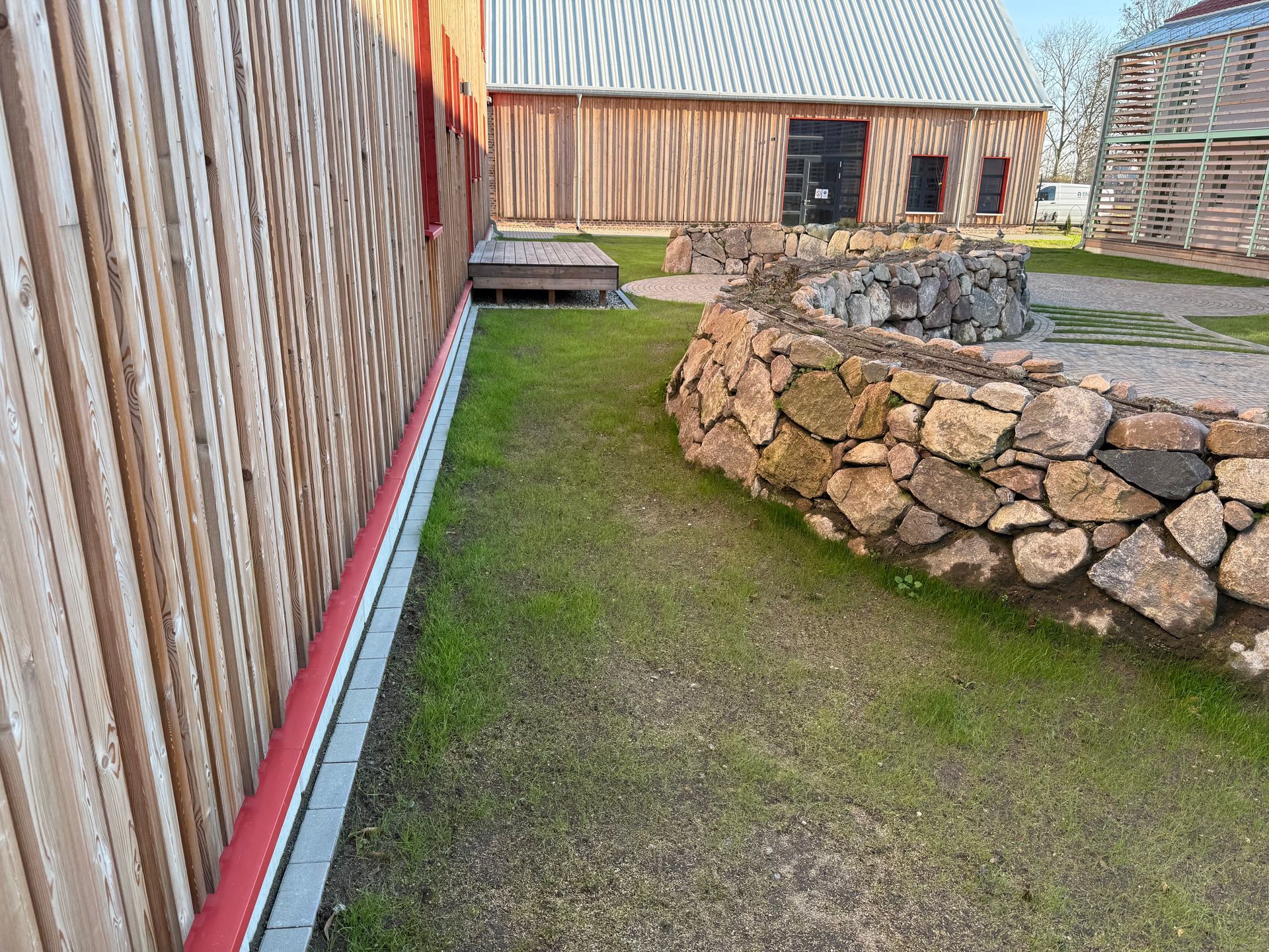 Äußeres Holzgebäude mit roter Dachrinne, grünem Gras und einer Steinmauer.