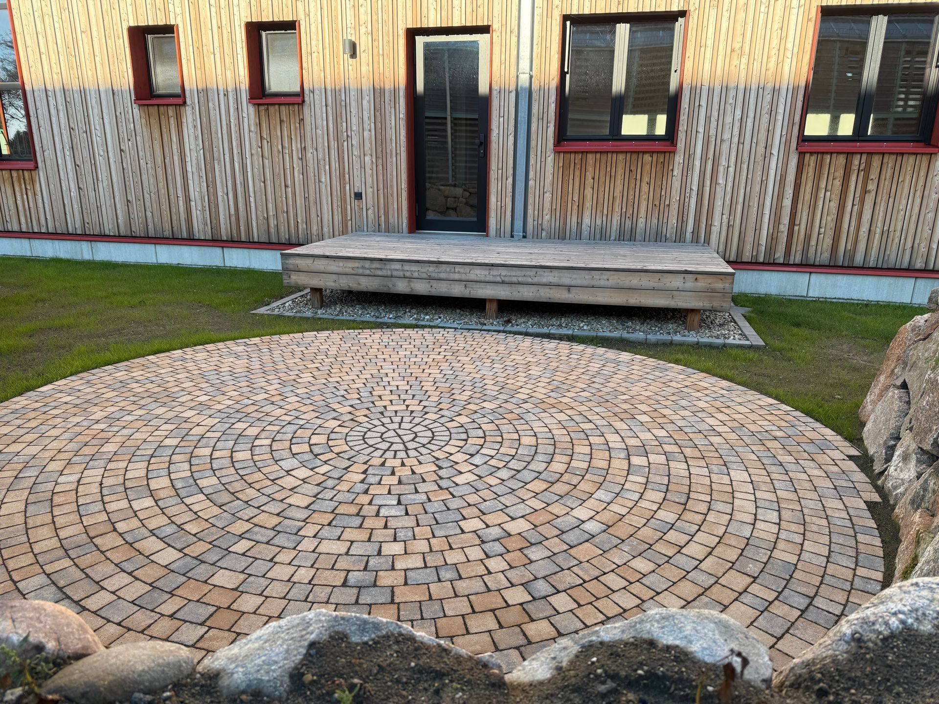 Runder Steinhof vor einer Holzterrasse und einem Gebäude mit Rasen und einer Steinumrandung.