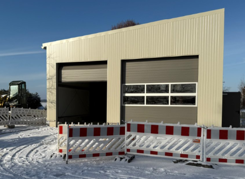 Ein beigefarbenes Metallgebäude mit zwei Garagentoren, eines davon offen. Rot-weiße Absperrungen im Schnee.
