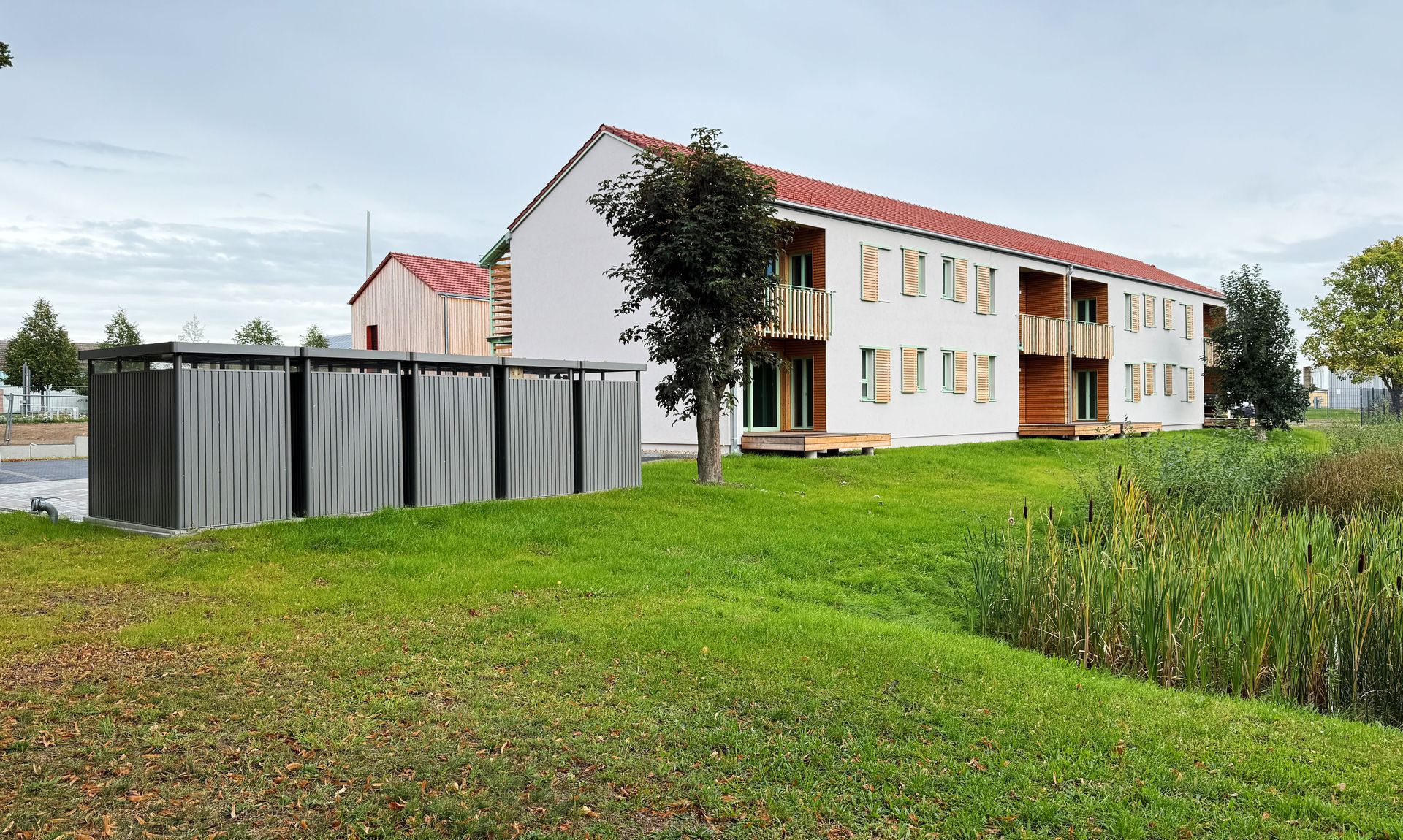 Zweistöckiges weißes Gebäude mit rotem Dach und Holzelementen. Graue Mülltonnen links, grüner Rasen.