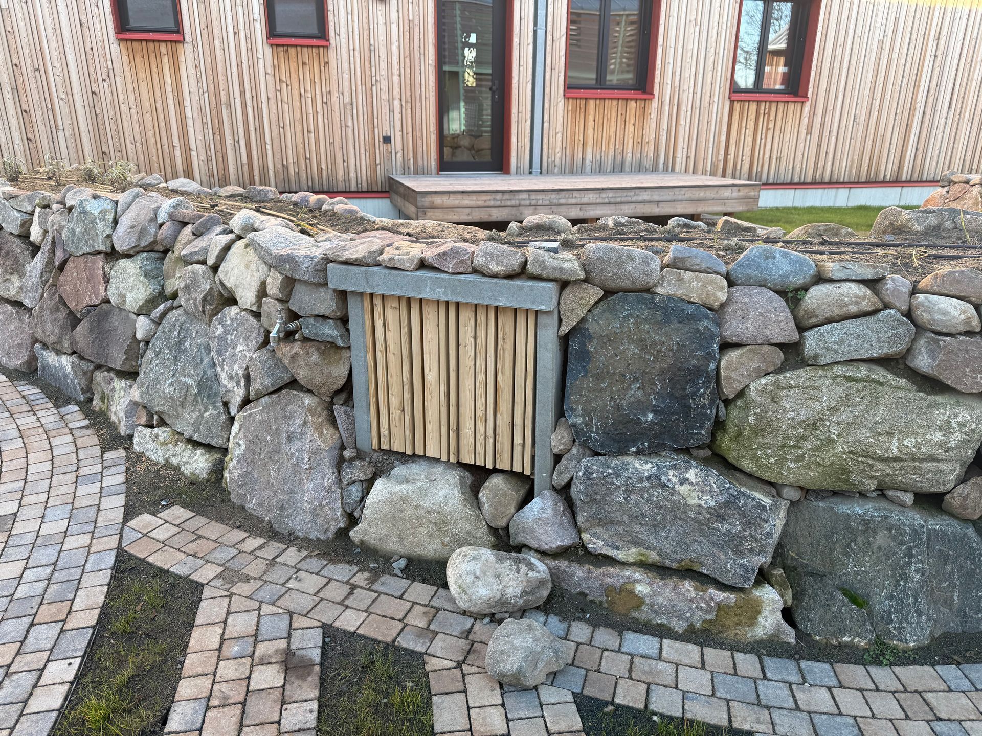 Steinmauer mit einem darin eingebauten hölzernen Insektenhotel vor einem holzverkleideten Gebäude.