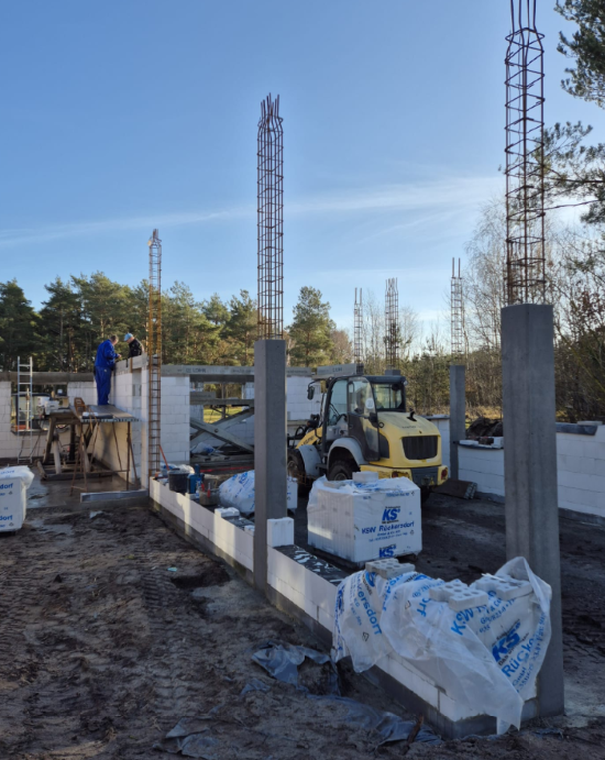 Baustelle mit Betonpfeilern und einem teilweise errichteten Gebäude. Ein Arbeiter und ein Traktor sind zu sehen.