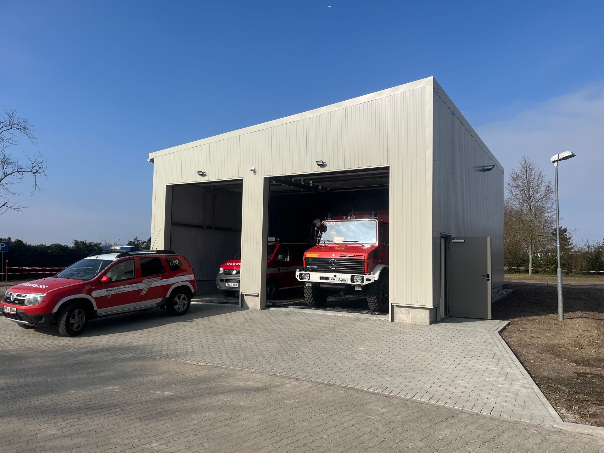 Feuerwache mit roten Feuerwehrwagen, die innerhalb und außerhalb des Gebäudes auf einem gepflasterten Platz geparkt sind.