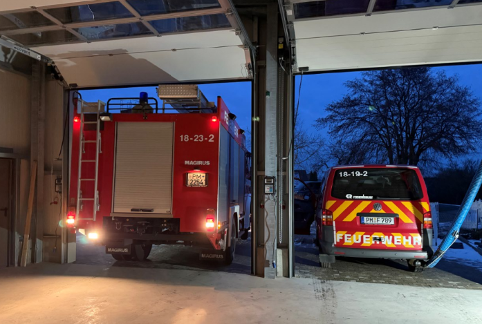 Feuerwehrwagen in einer Garage mit offenen Toren. Rote und weiße Fahrzeuge vor dem Abendhimmel.
