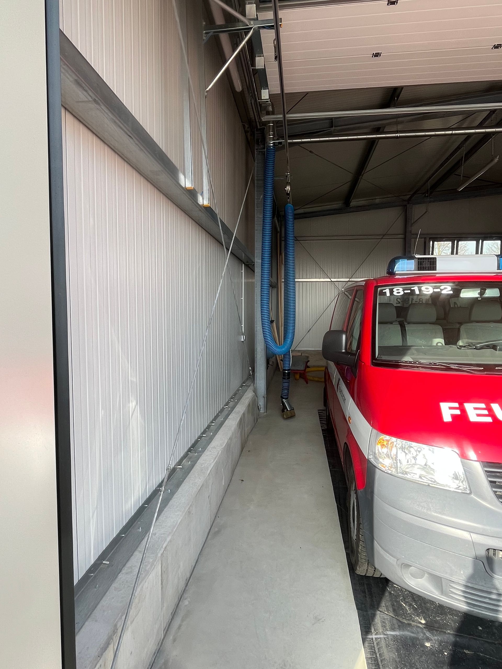 Feuerwehrwagen in einer Fahrzeughalle; blauer Schlauch hängt von der Decke. Links weiße Wellblechwand.