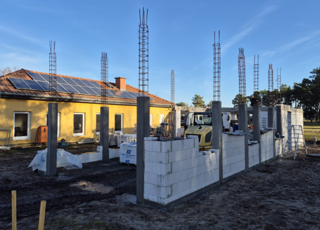 Baustelle mit teilweise errichteten Blocksteinmauern, Betonpfeilern und einem bestehenden gelben Gebäude mit Solarpaneelen.