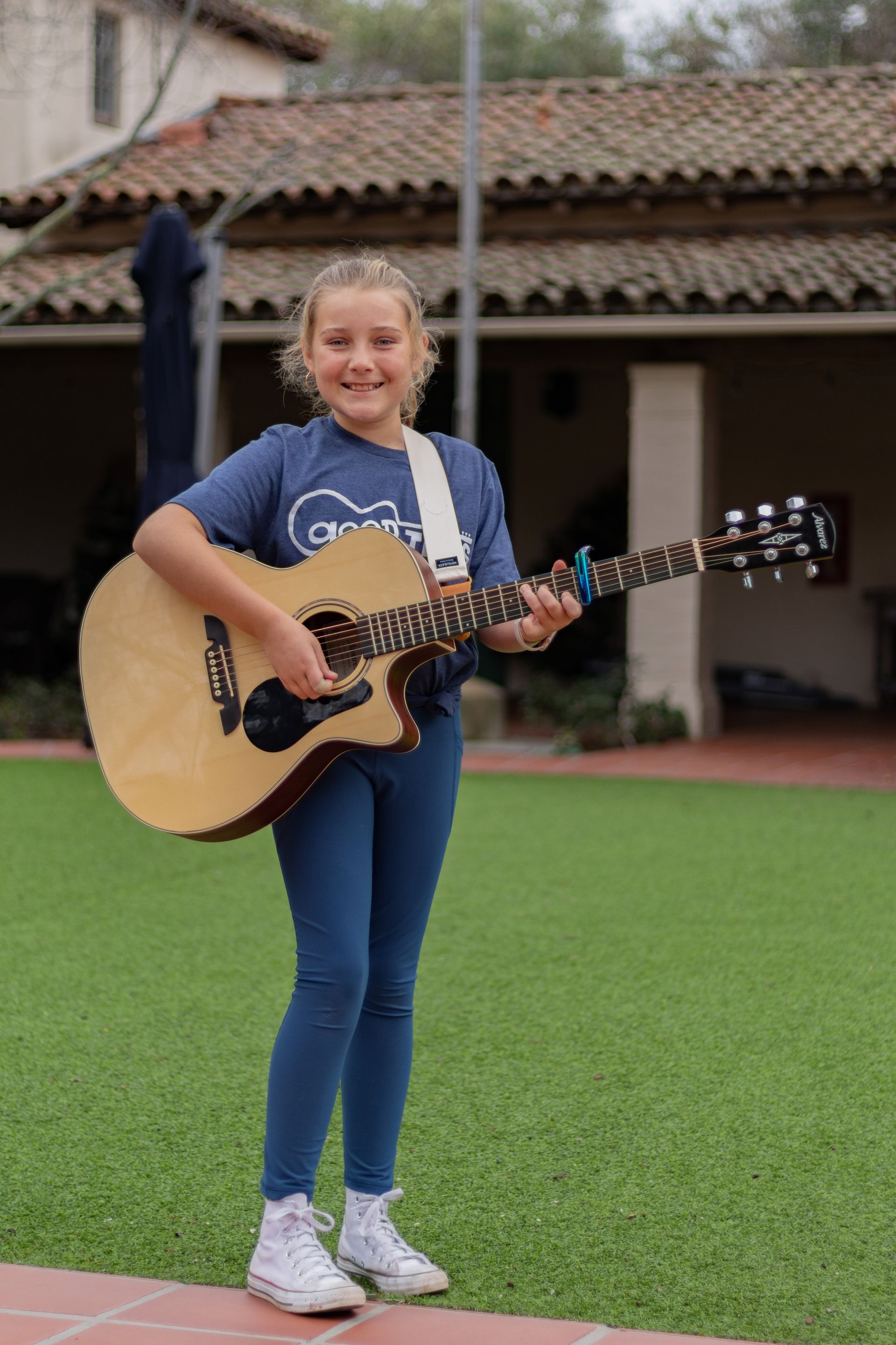 Girl playing acoustic guitar outdoors, smiling. She is wearing a blue Good Times Guitar t-shirt.