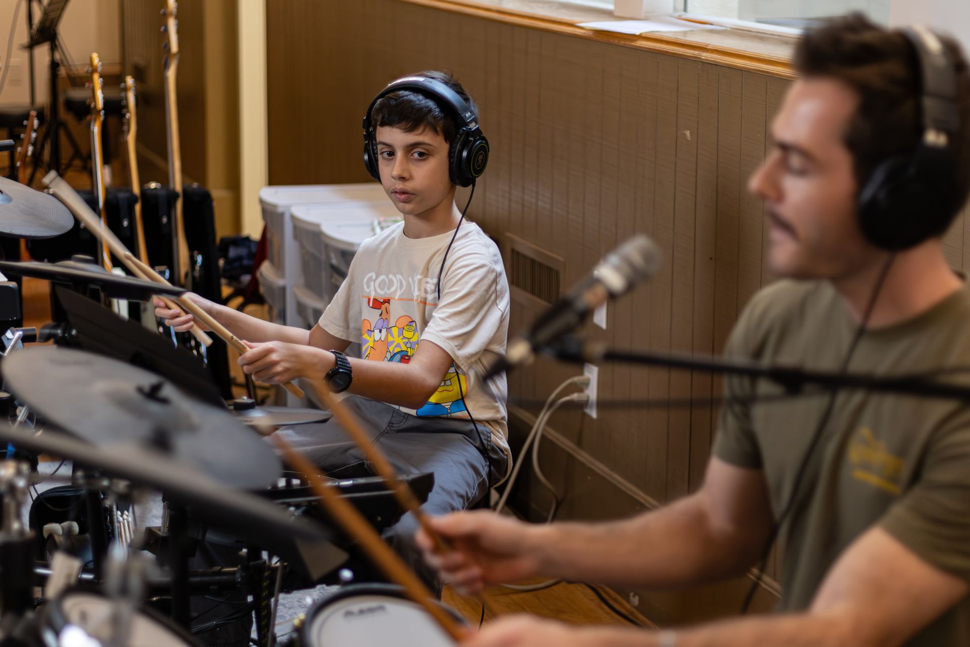 Music student and instructor playing drums in a studio. Both wear headphones.