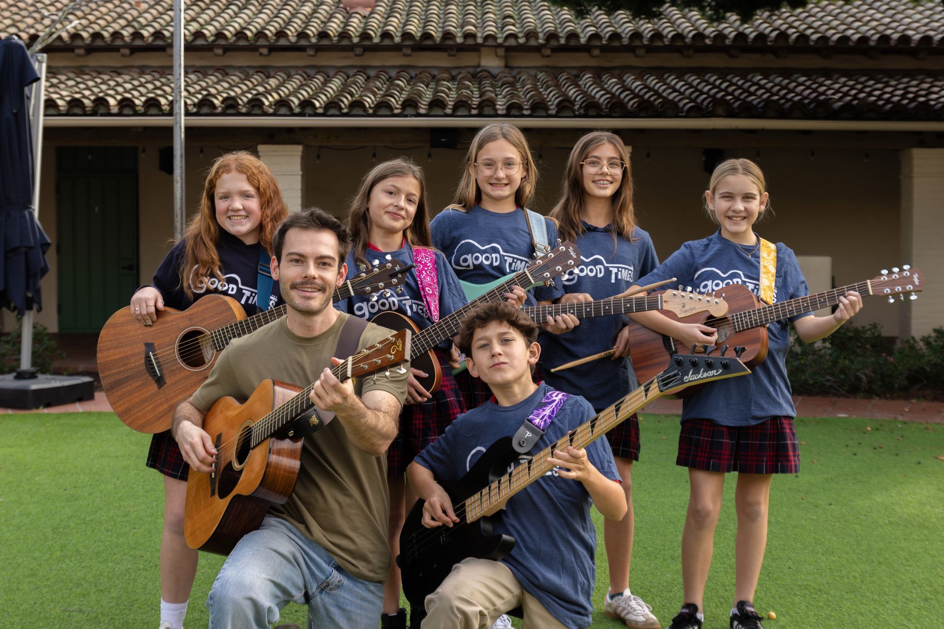 A group of children and their music teacher with guitars pose on a lawn. 