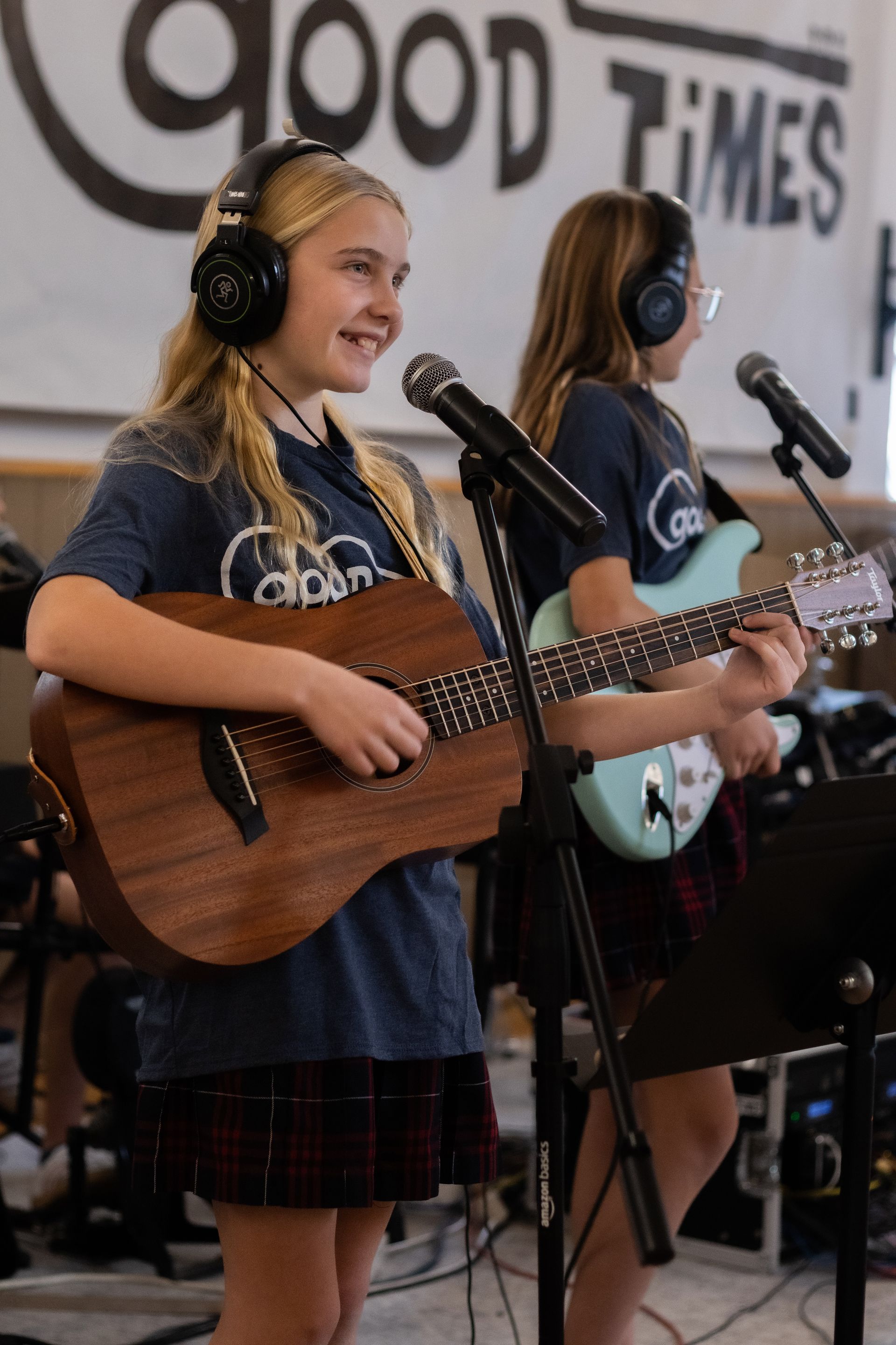 Two girls playing guitars and singing into microphones in a band setting. One wears headphones and smiles.