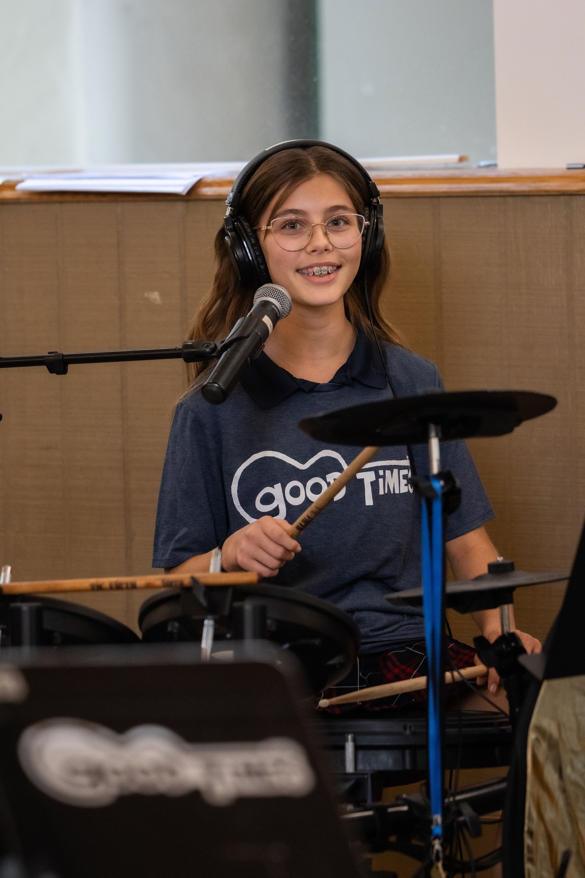 Young teen wearing headphones playing electronic drums, smiling in a room with a microphone.