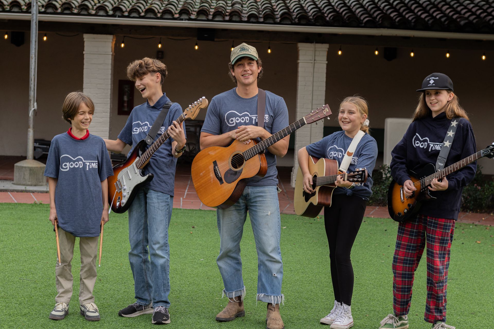 Four students holding their guitars and drum sticks posing and smiling outside with their music teacher.