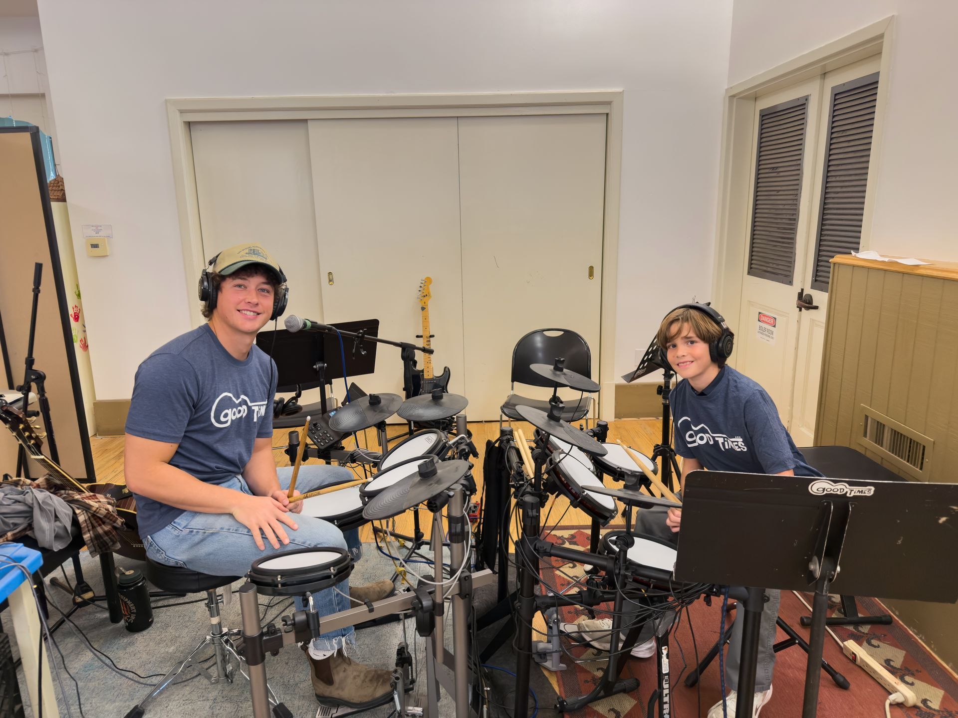A drum student and his teacher wearing headphones seated at electronic drum sets in a room; smiling.