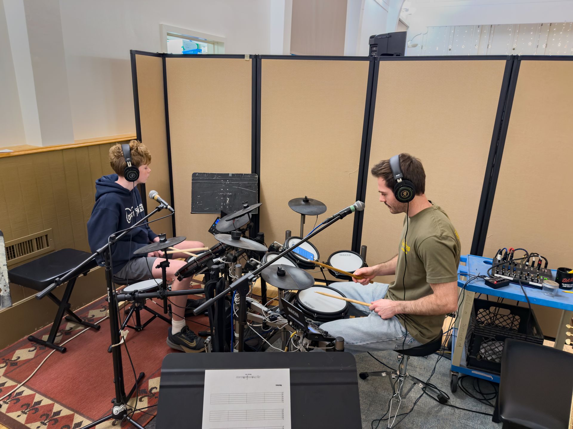 A student and teacher playing electronic drums in a room. The student is speaking into a microphone.