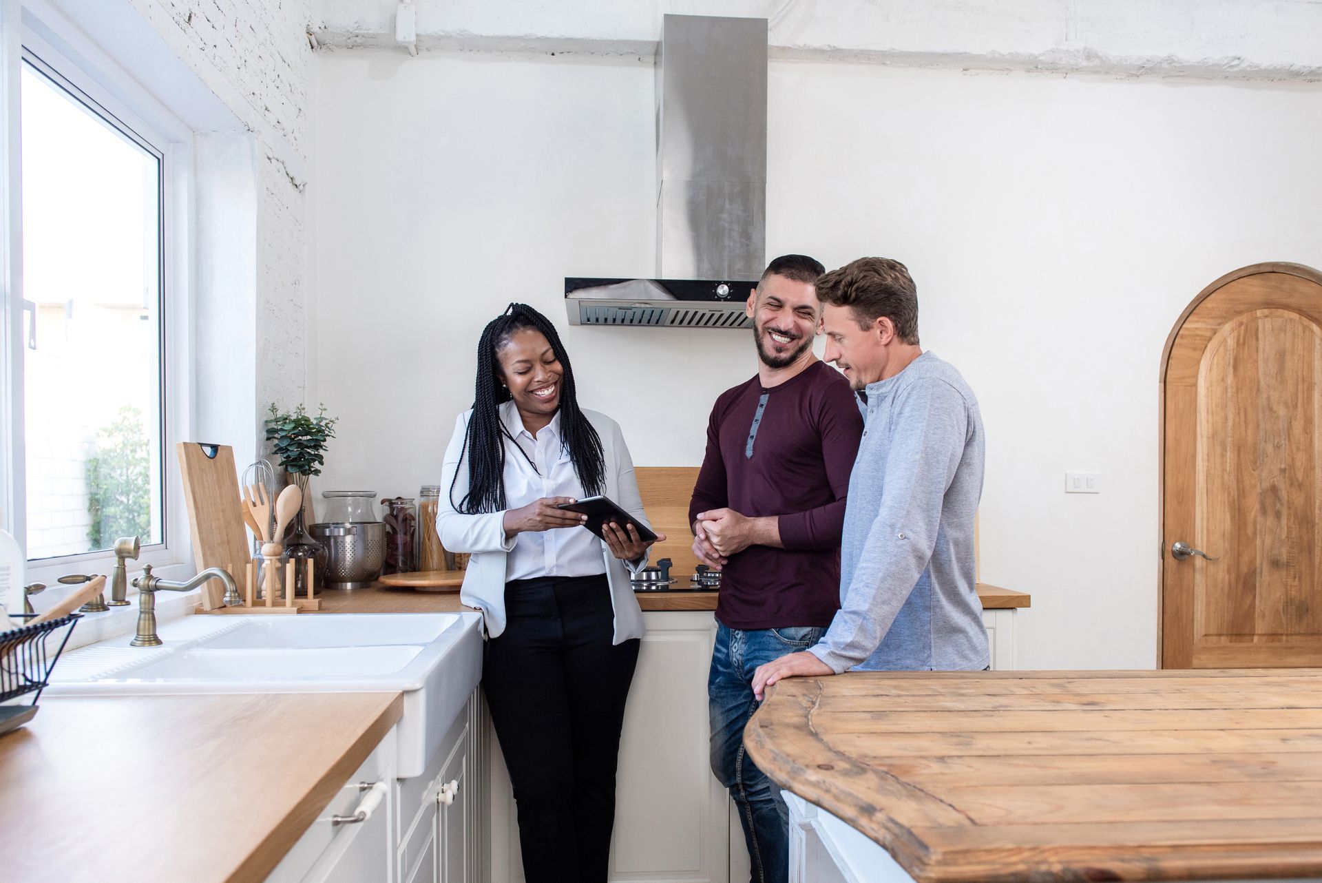 Realtor shows tablet to smiling same-sex couple in a bright kitchen; one leans on counter.