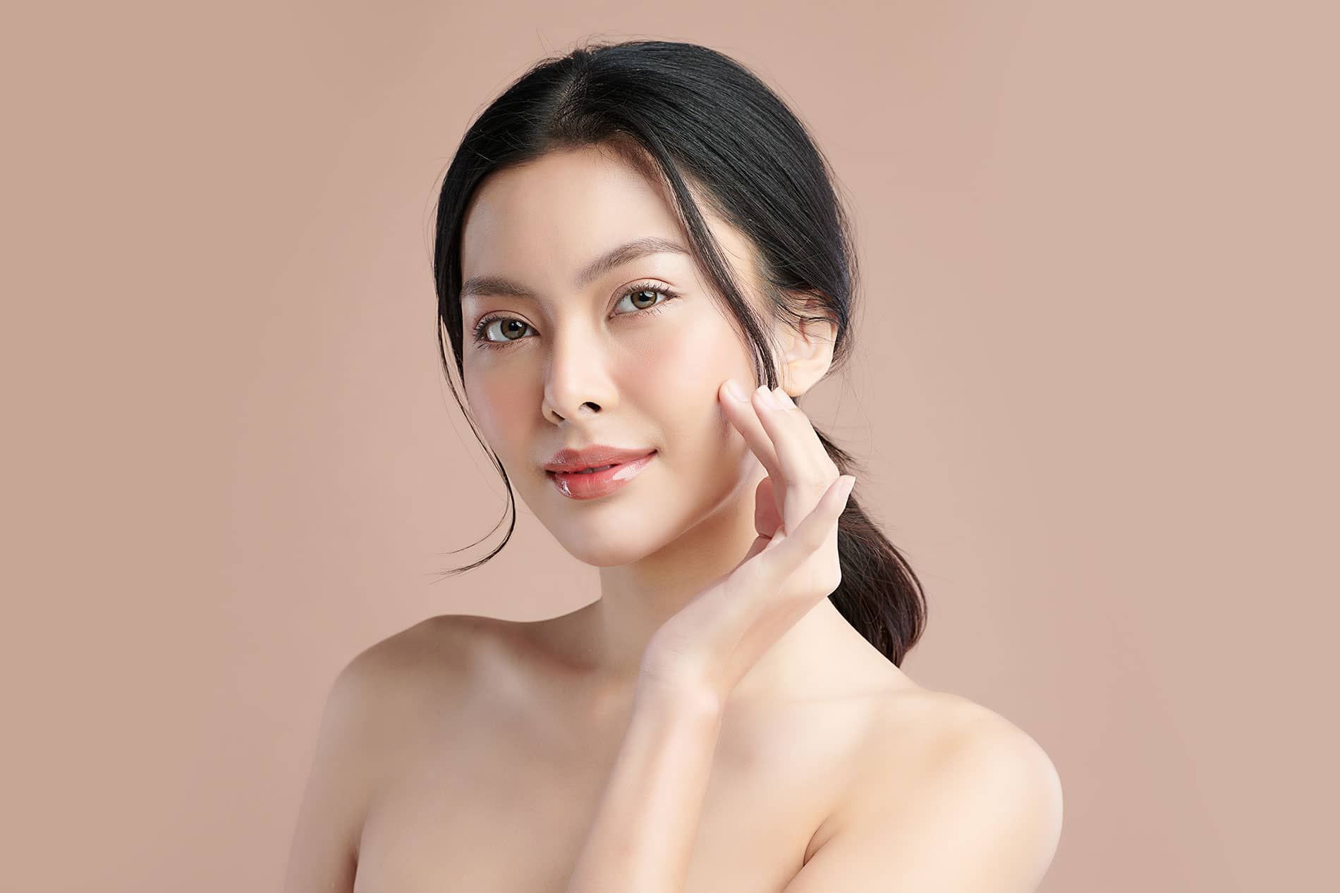 A young woman with dark hair and a bare shoulder gently touches her cheek, smiling softly against a neutral background.