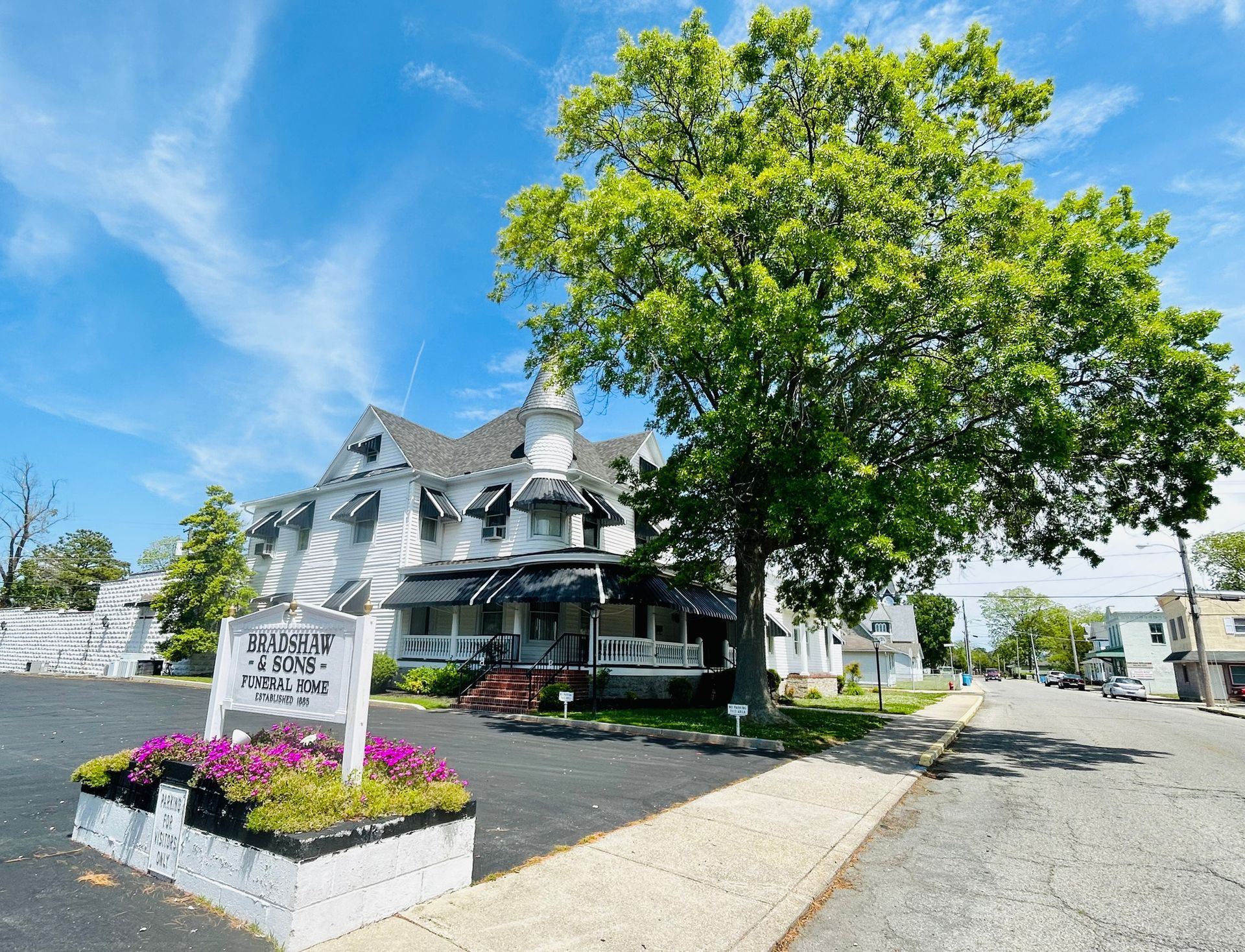 White Victorian building with black awnings, sign, and flowering plants. Large tree on right.