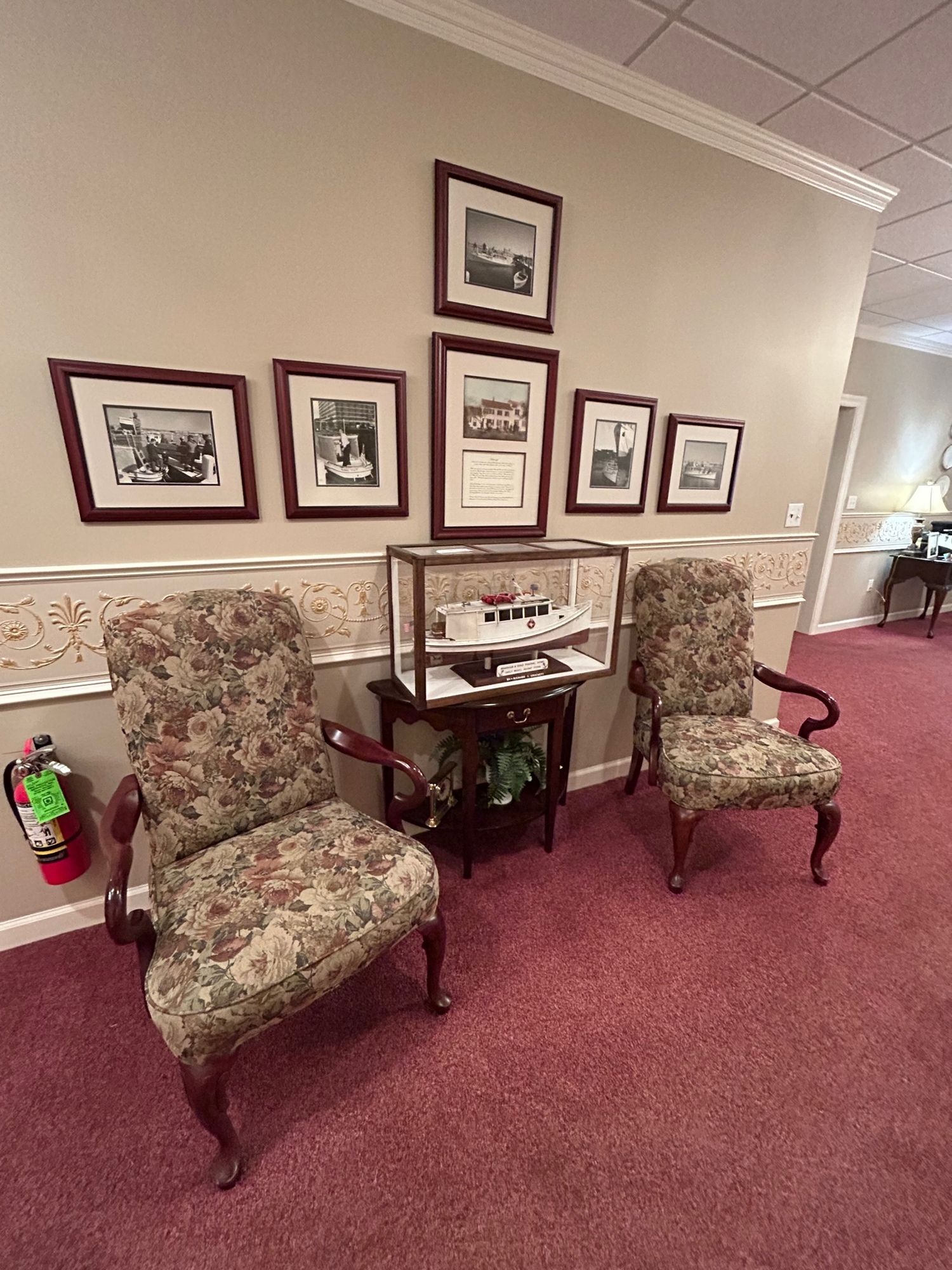 Two floral armchairs, display case with a ship model, framed photos, and a fire extinguisher against a pink-carpeted wall.