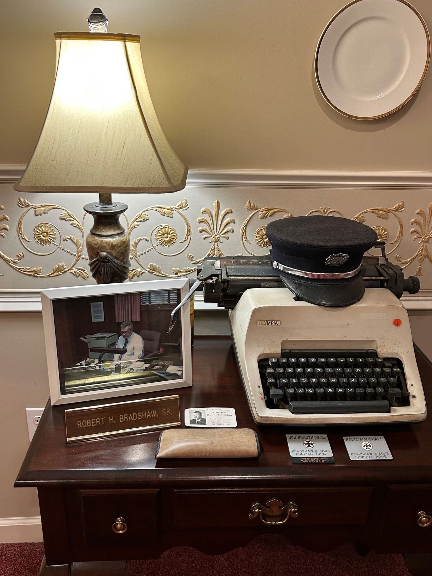Desk with typewriter, lamp, cap, photo, cards, and decorative wall.