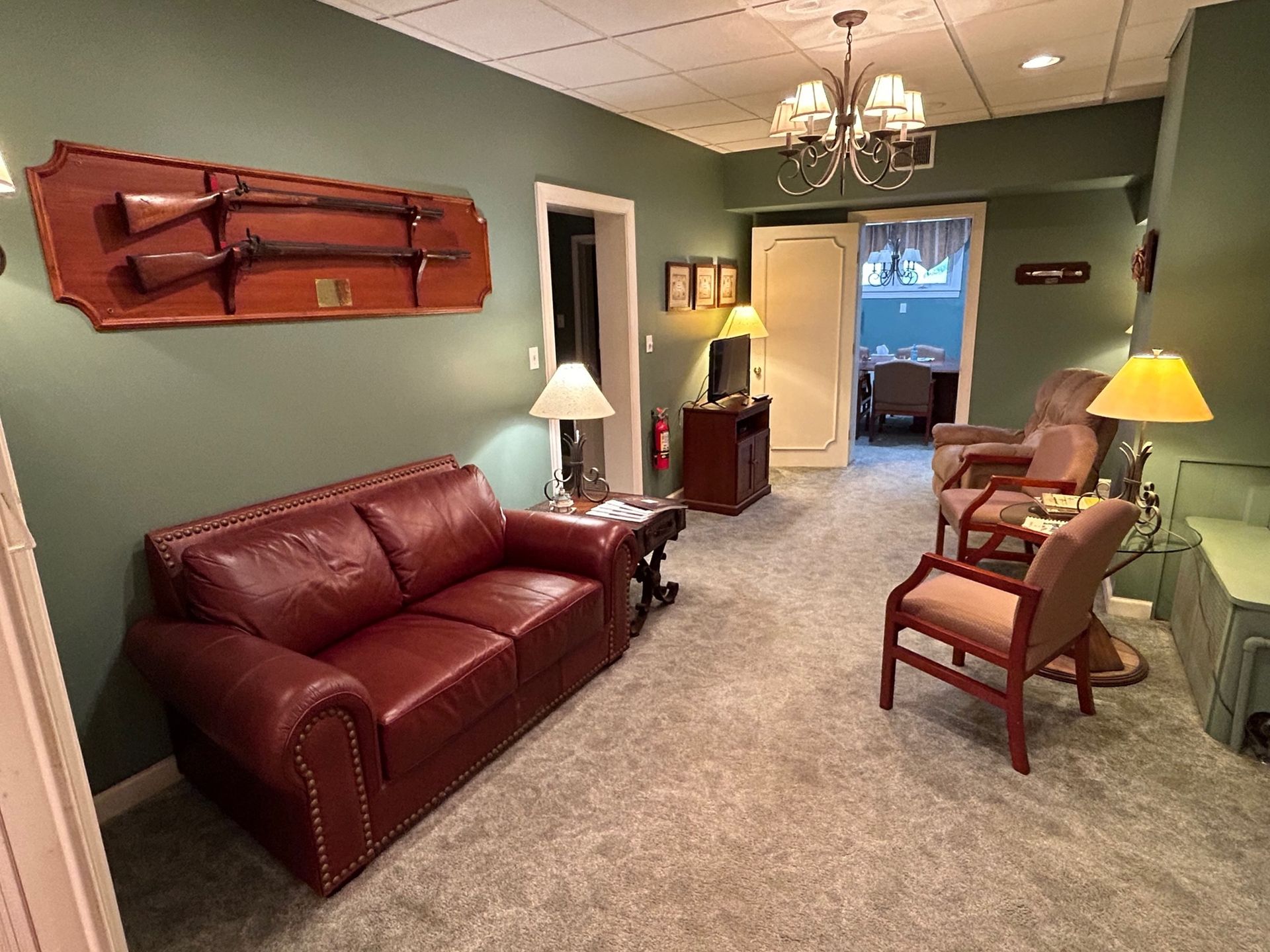 A room with a red leather couch, green walls, and a mounted rifle display. There's a chandelier and seating area.