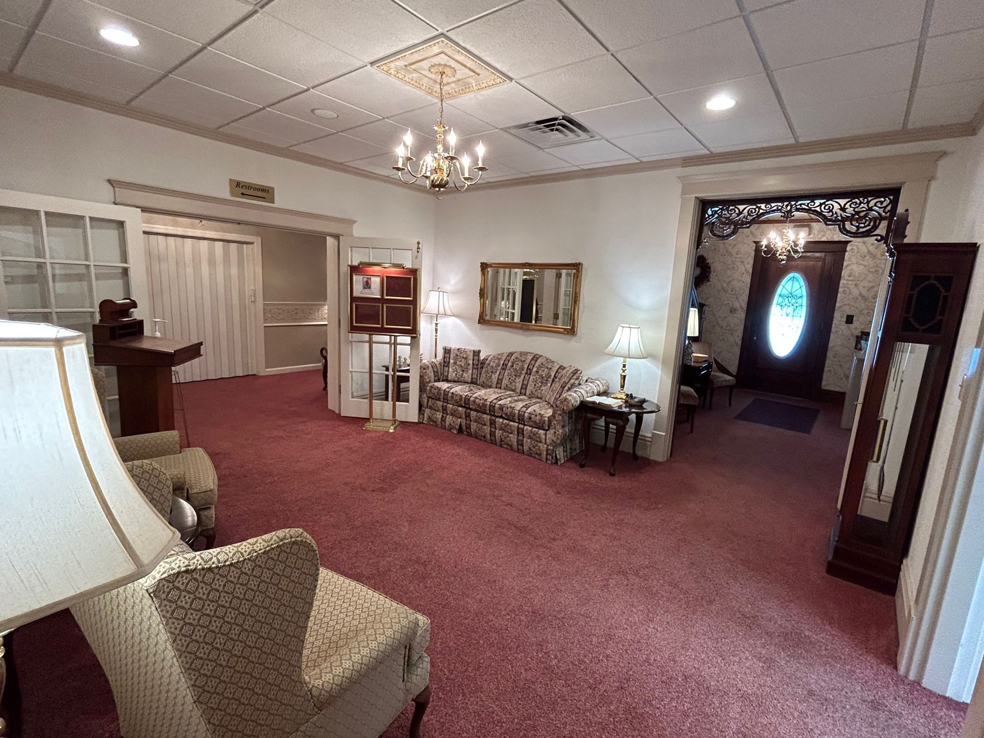 Interior of a room with floral sofa, chairs, a mirror, and a grandfather clock on a pink carpet.