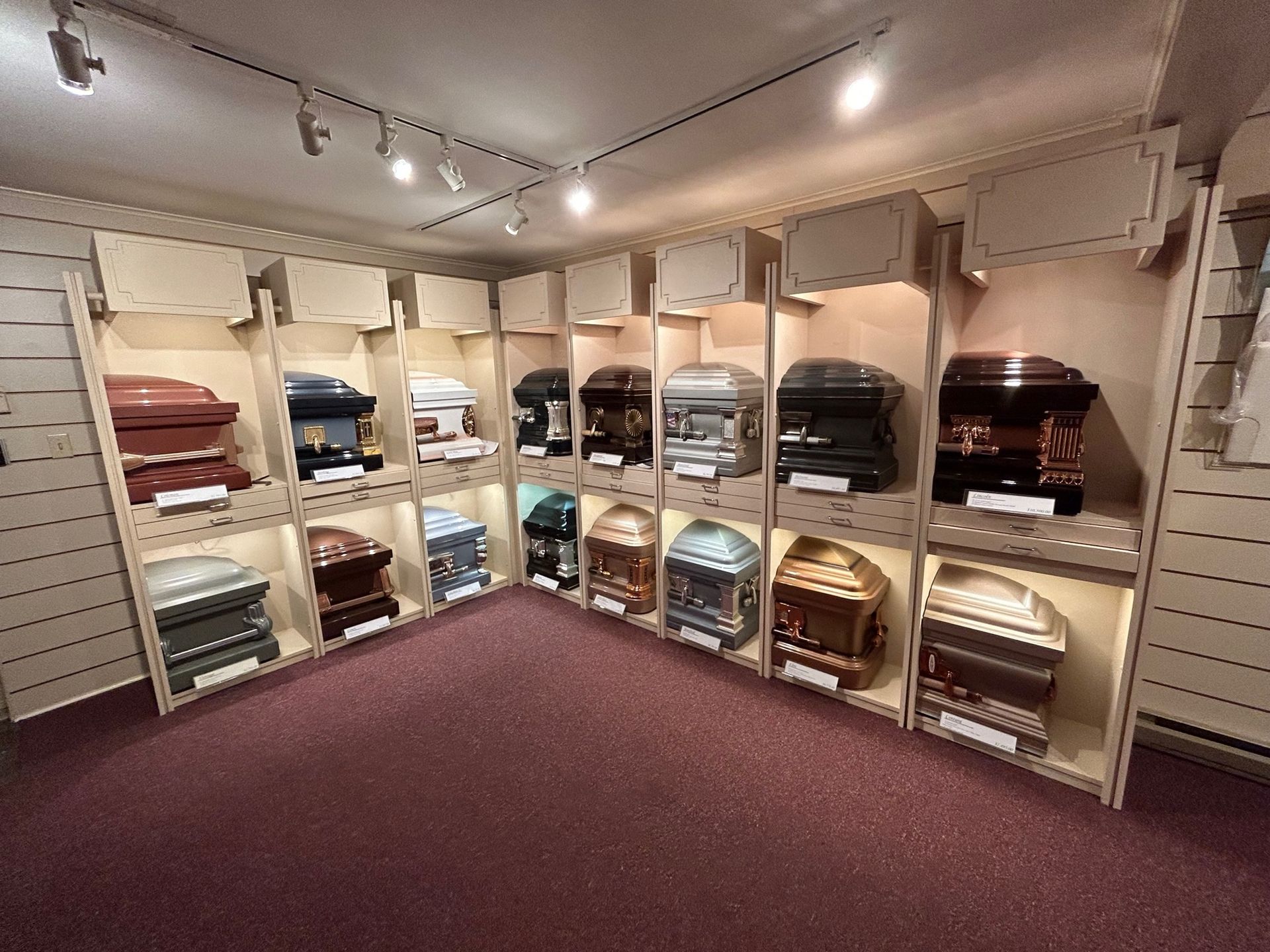 Display of caskets in a brightly lit showroom with a maroon carpet. Various colors and styles are visible.
