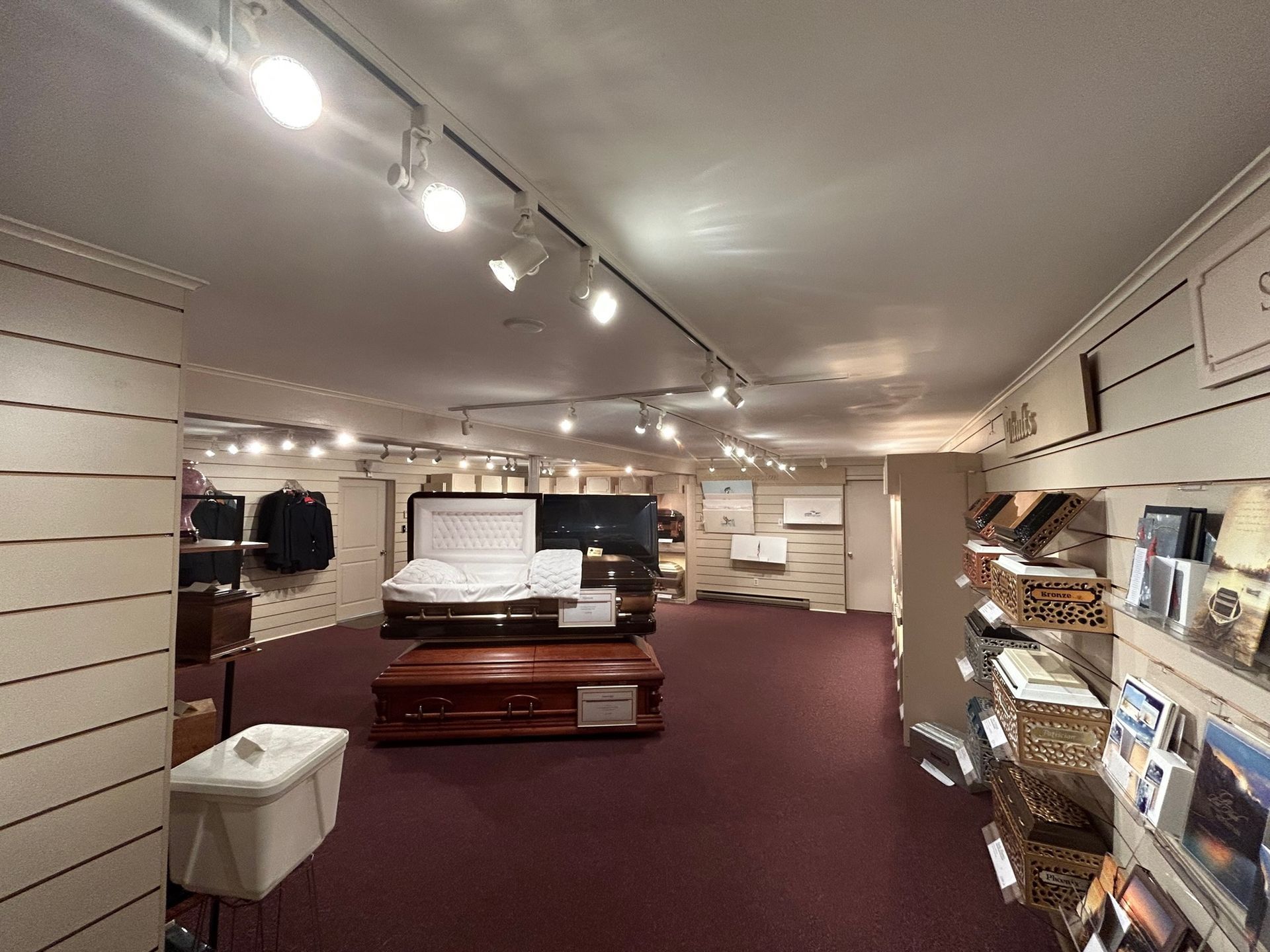 Interior of a funeral home displaying caskets, urns, and memorial cards. Red carpet, white walls, and track lighting.