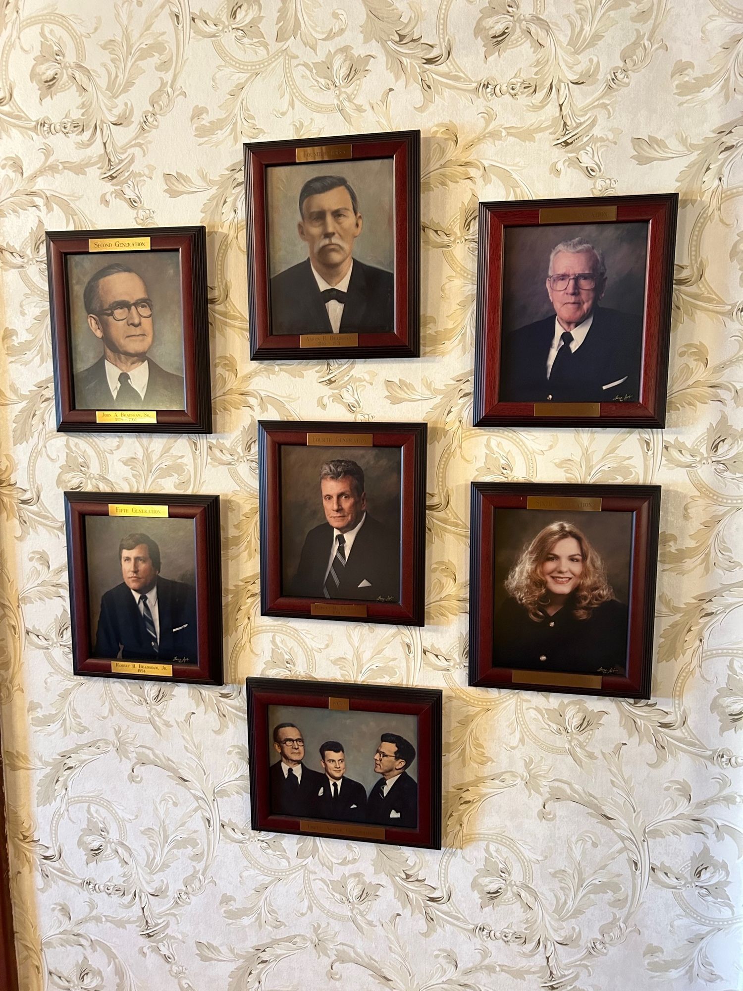 Portraits in ornate wooden frames hang on patterned wallpaper, featuring individuals in formal attire.
