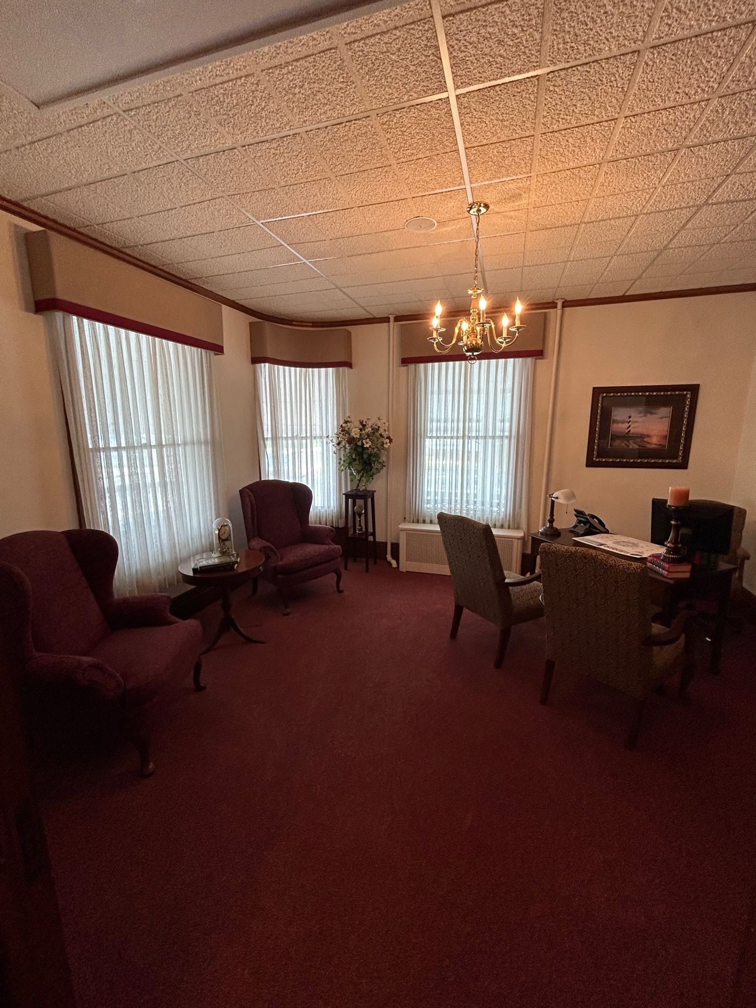 Office with red carpet, chairs, and desk. Windows with curtains and a chandelier.