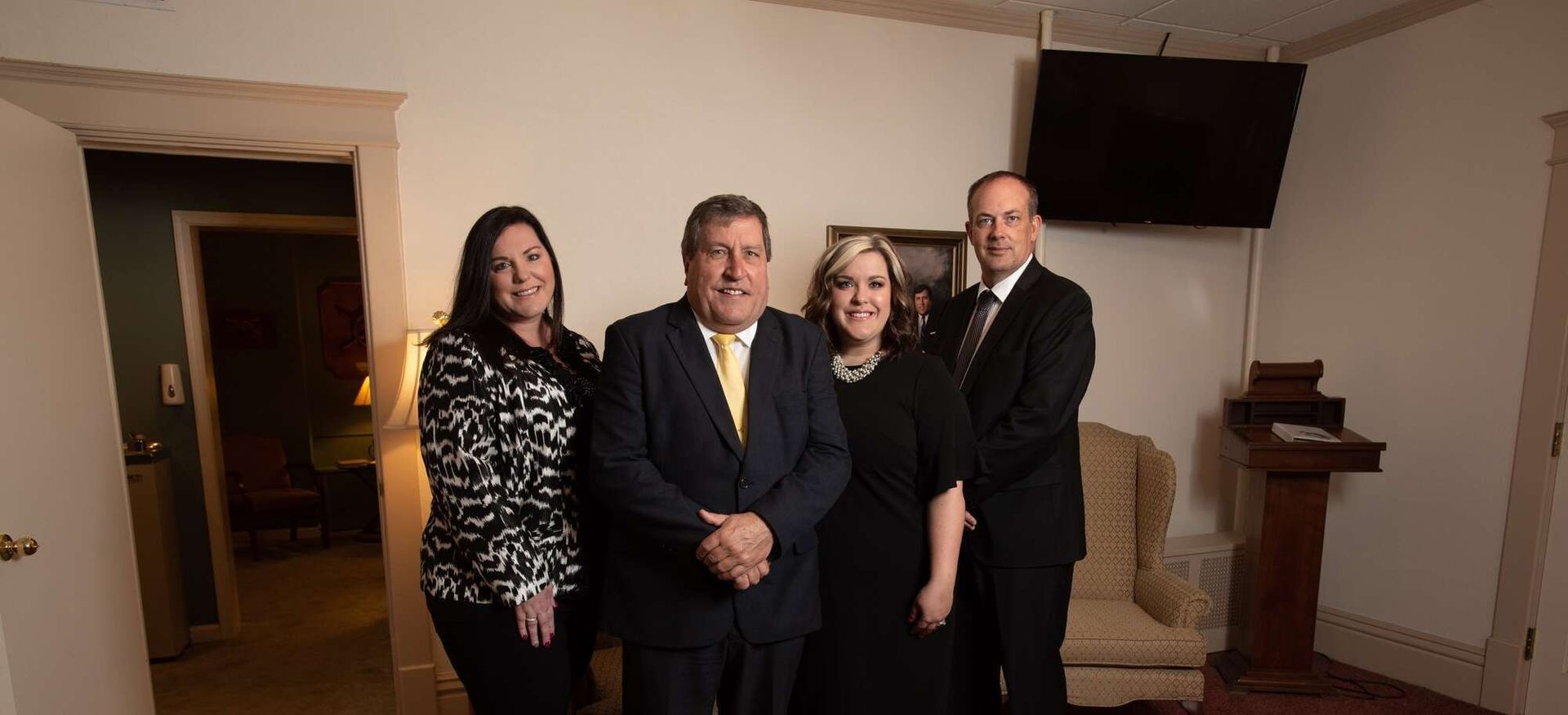 Four people standing in a room, two women, and two men. One man in a navy suit with a yellow tie, the others in black.