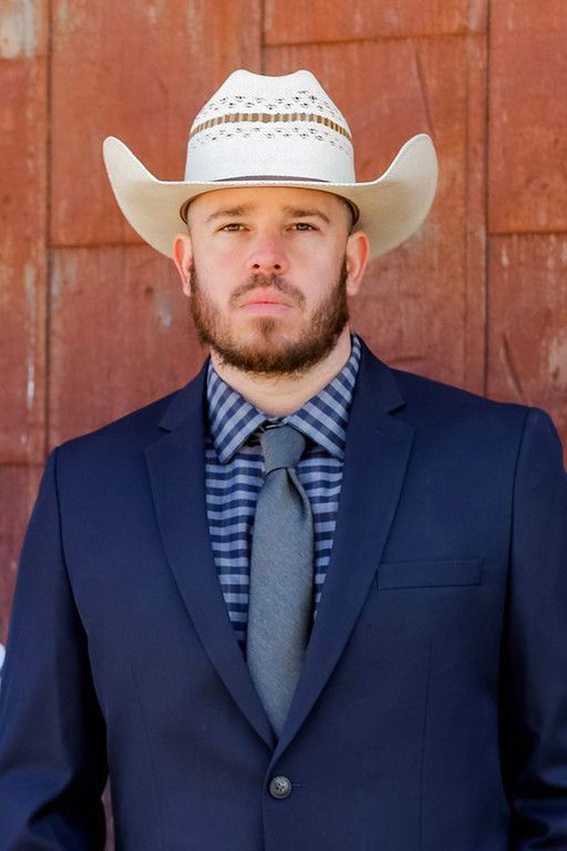 A man in a suit and cowboy hat is standing in front of a red wall.