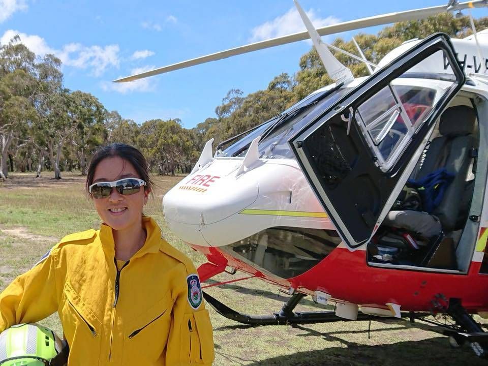 Woman in yellow flight suit stands beside a red and white helicopter on a sunny day.