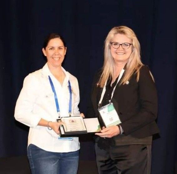 Two women at a conference; one presents a plaque to the other, both smiling.