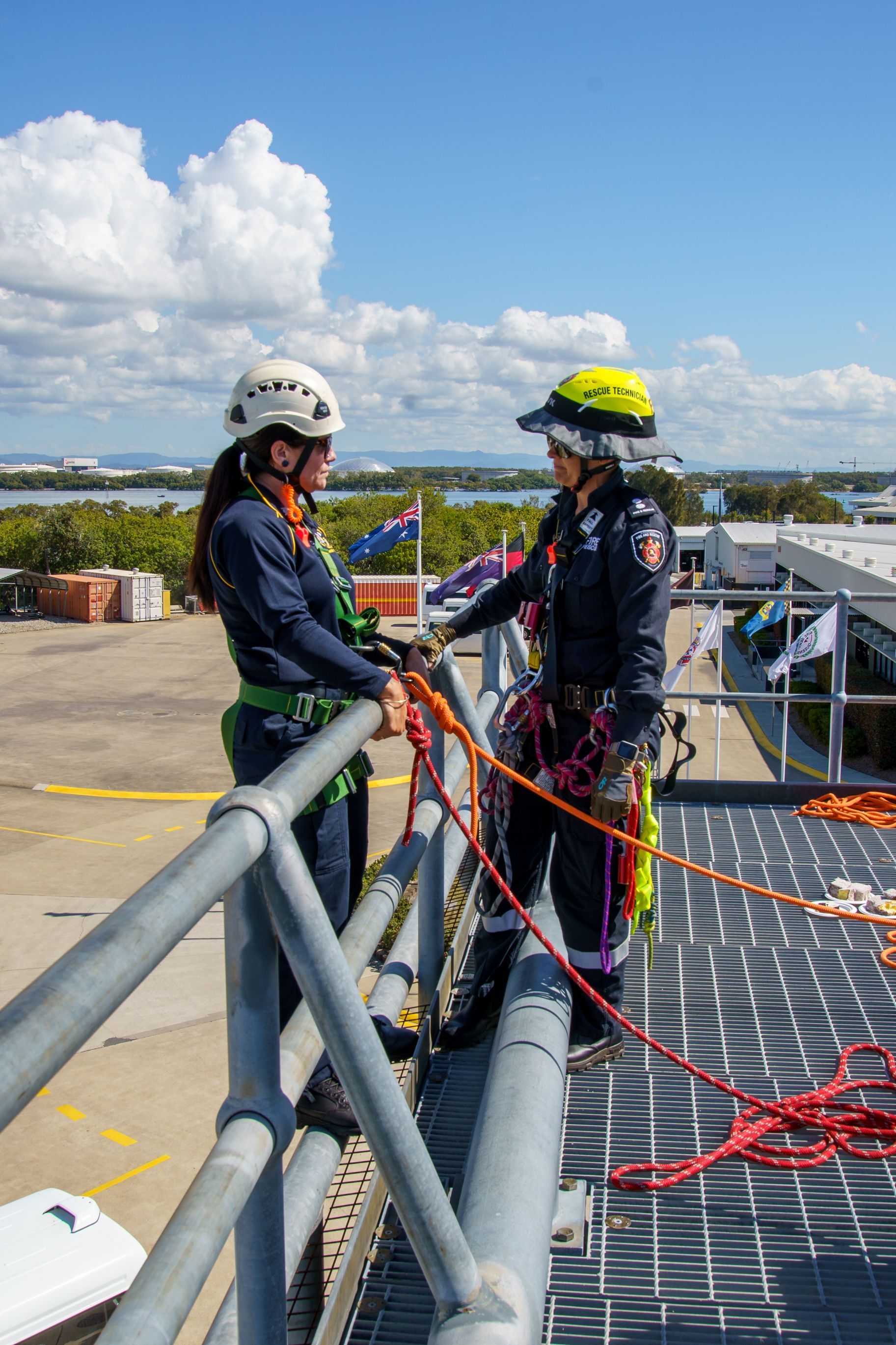 Two people in safety gear on a rooftop, discussing ropes and equipment. Sunny, blue sky in the background.