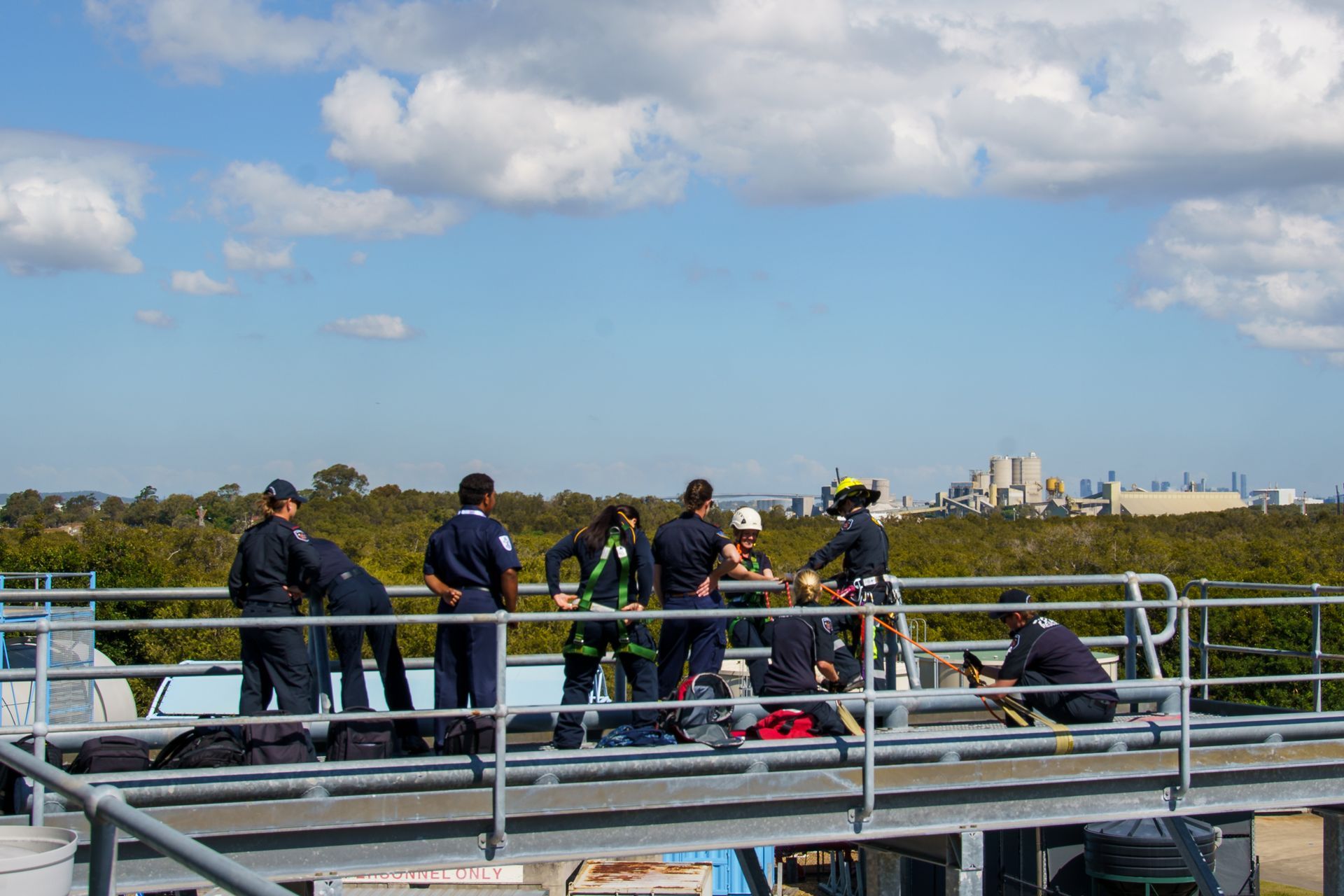 Firefighters on a rooftop; some in harnesses. Clear, sunny day; a cityscape in the distance.