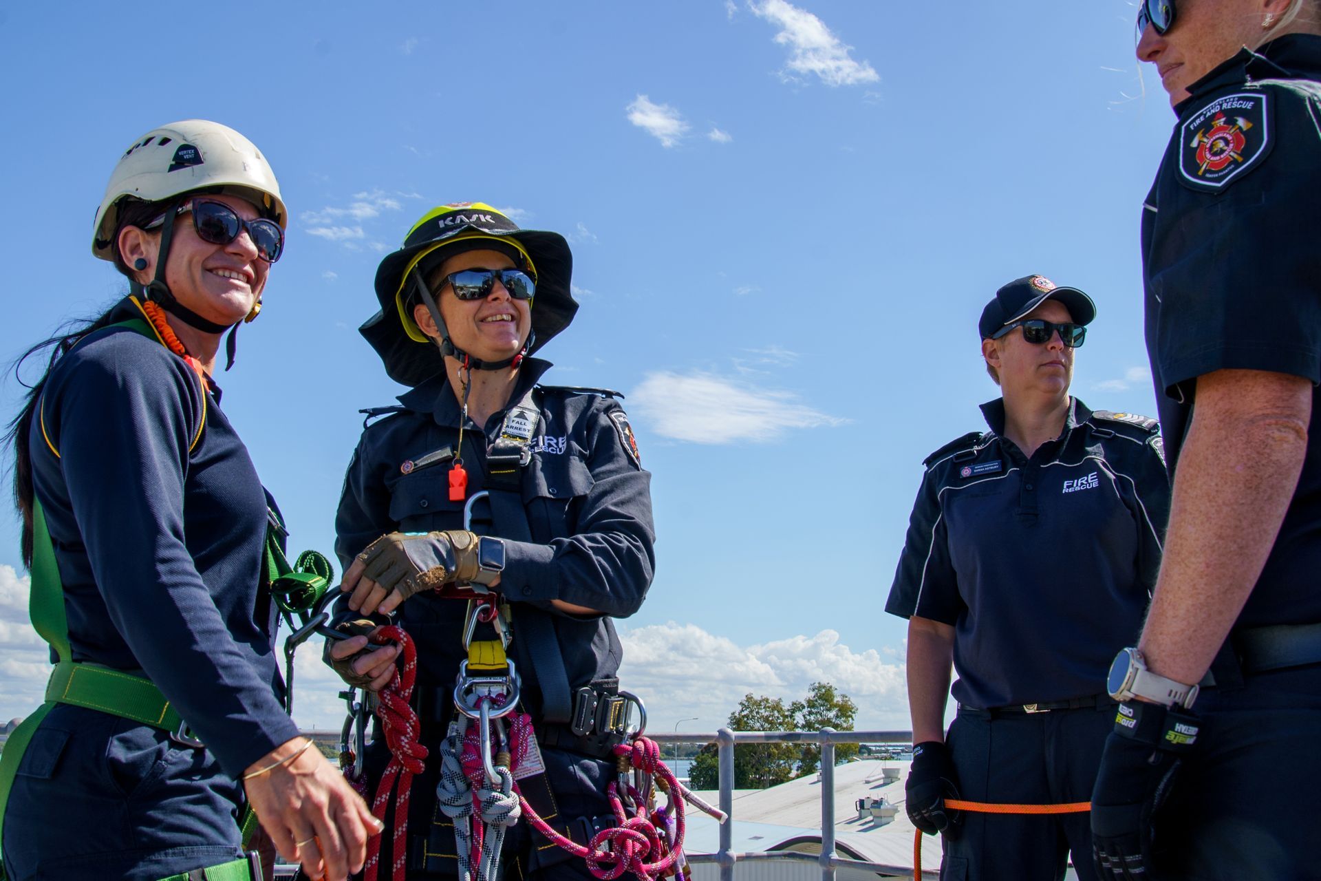 Four people in rescue gear on a rooftop; two women are smiling, blue sky background.