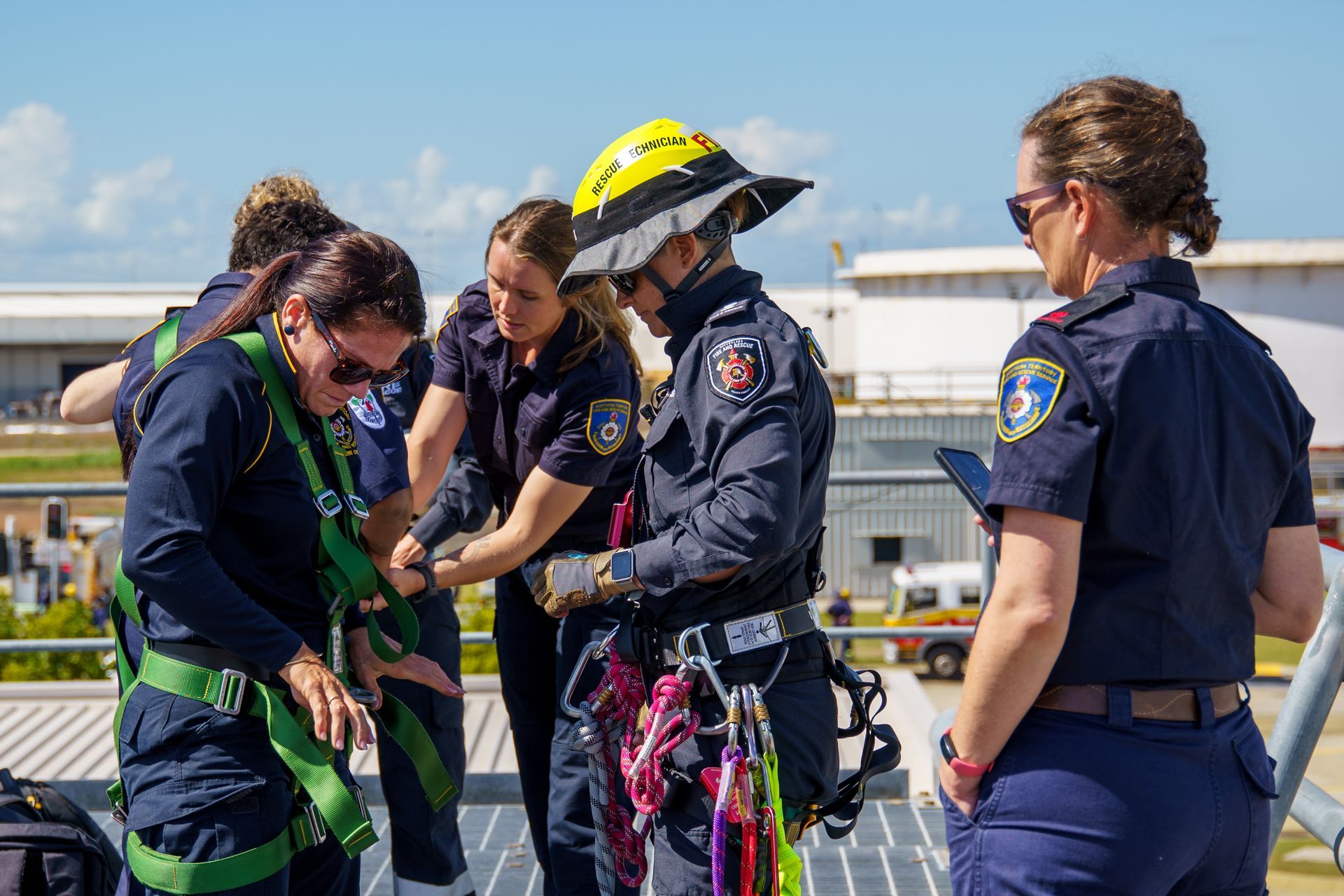 Firefighters in dark uniforms, practicing rope rescue on a rooftop. Yellow helmet, safety harnesses, checking equipment.