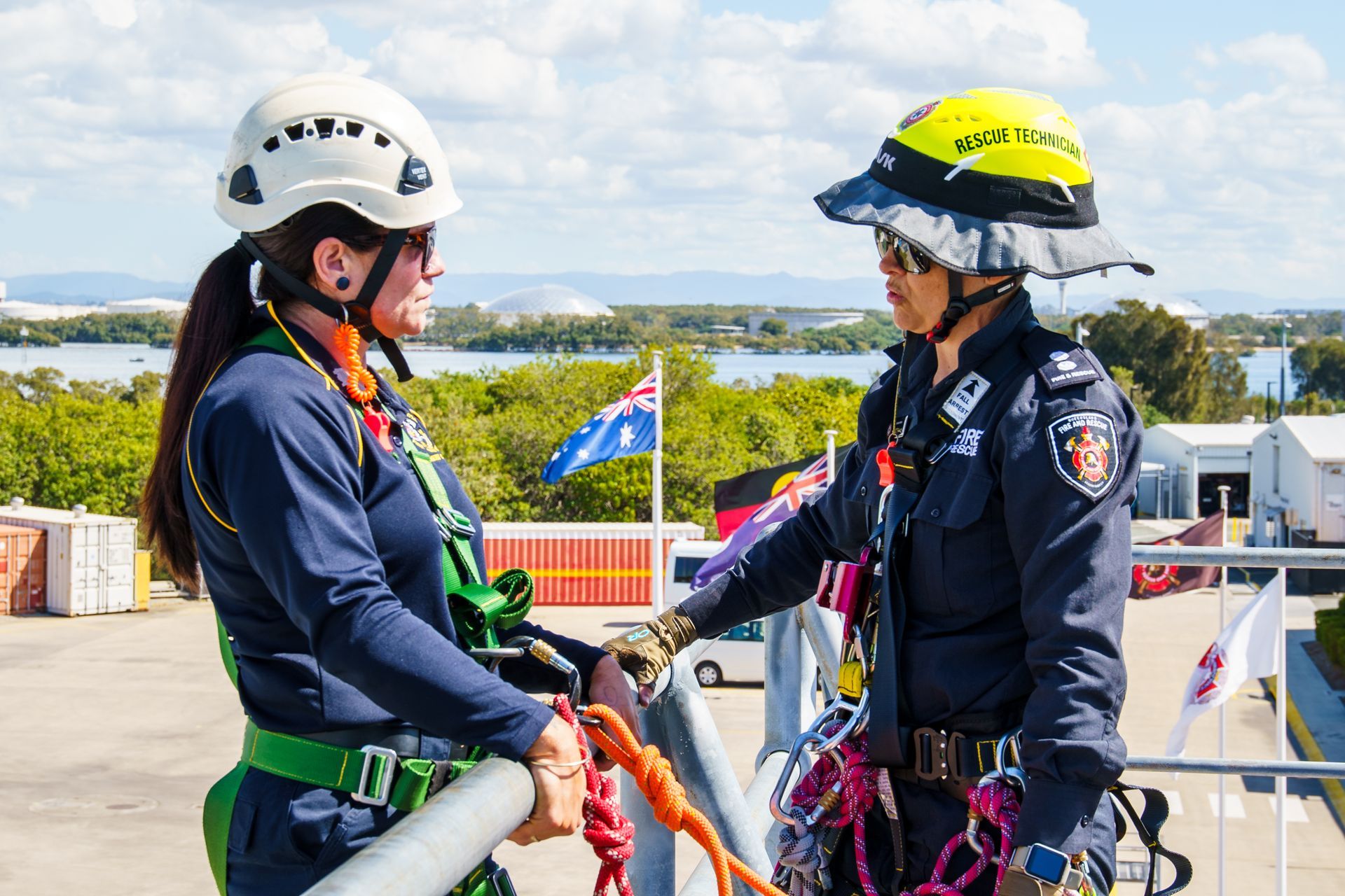 Two women in safety gear on a rooftop; one in a helmet and harness, the other in a police uniform, talking near an Australian flag.
