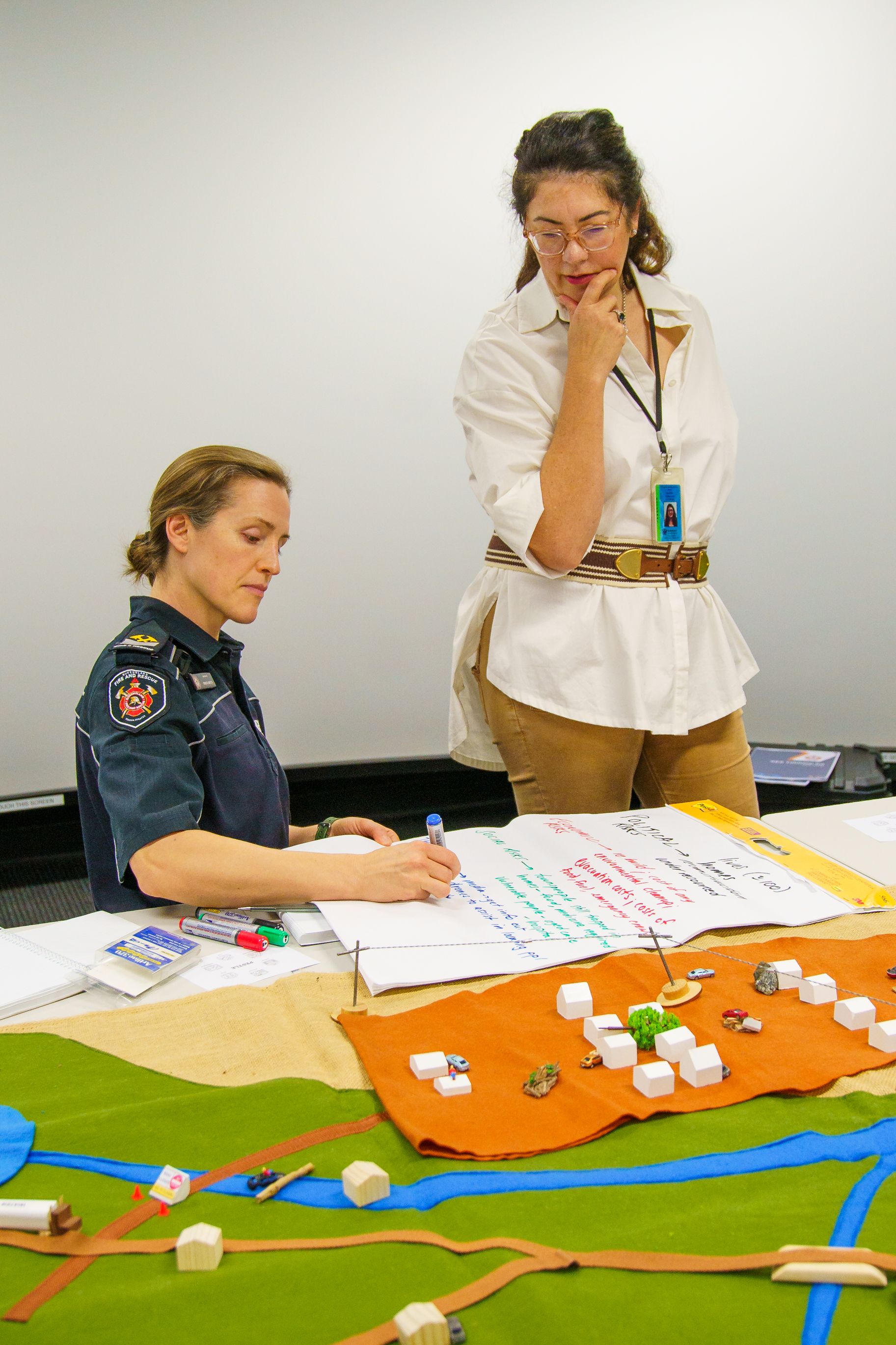 Two women looking at a map, one writing, the other thoughtful, in front of a projection screen.