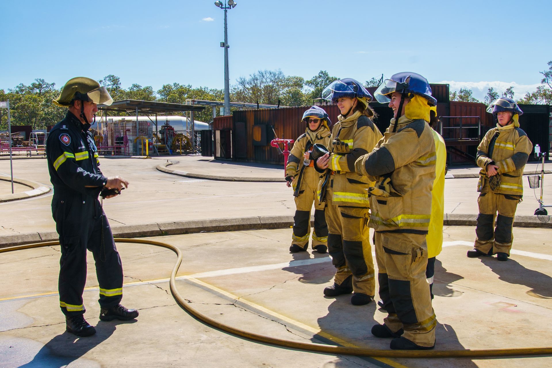 Firefighters in gear listen to an instructor on a training ground.