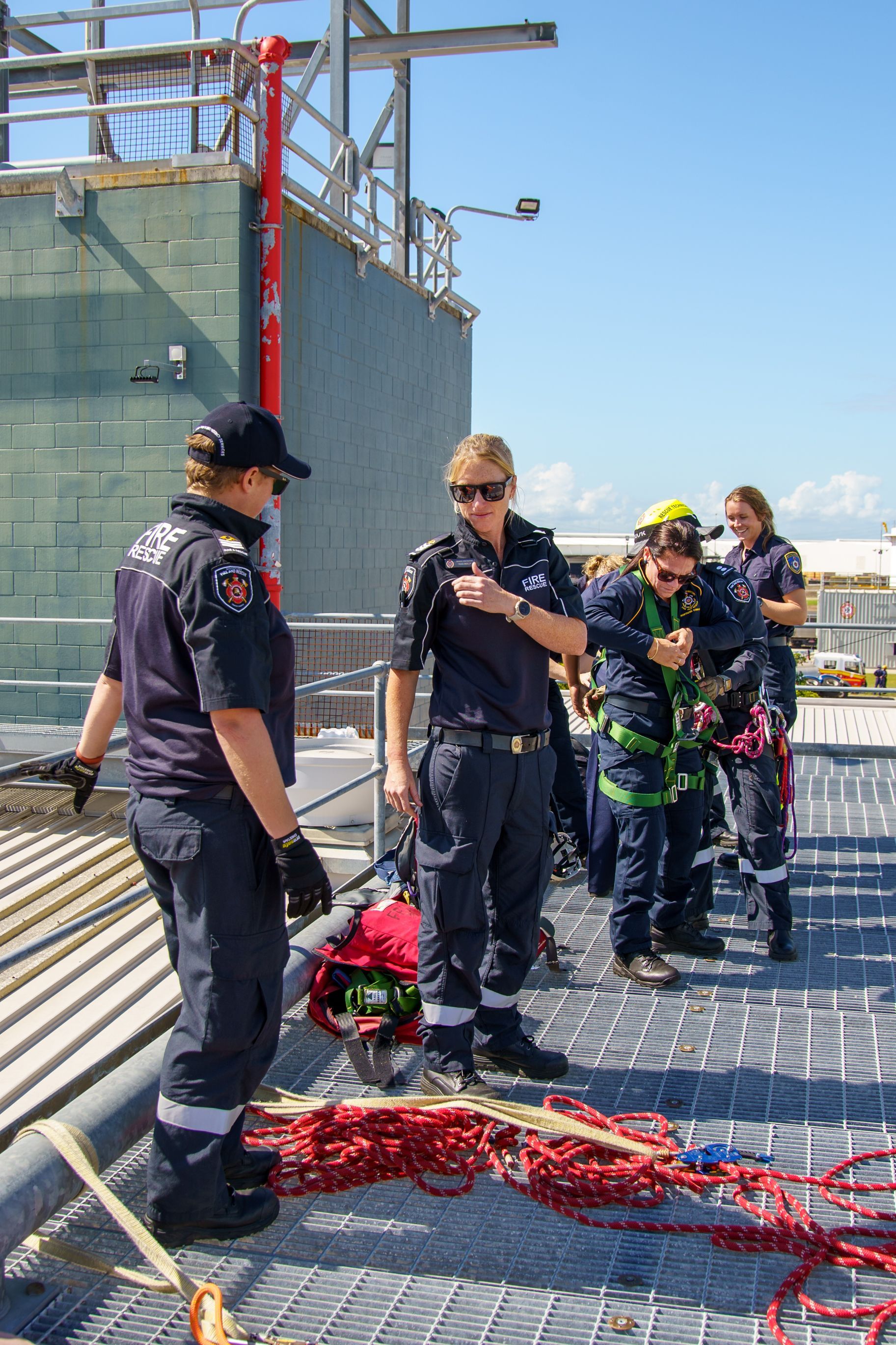 Firefighters on rooftop, discussing equipment. Two in uniform, two in harnesses; bright sky.