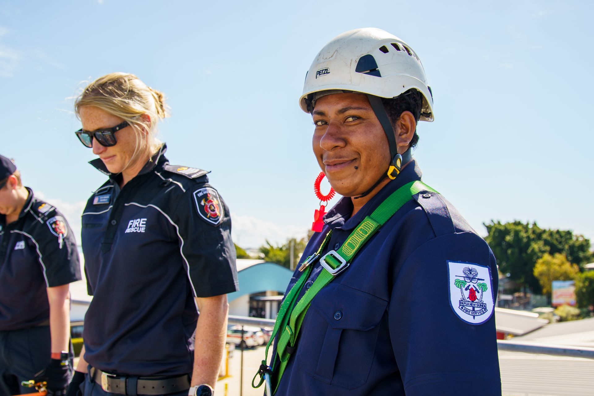 Firefighter in uniform wearing a harness and helmet smiles outdoors; another firefighter looks on.
