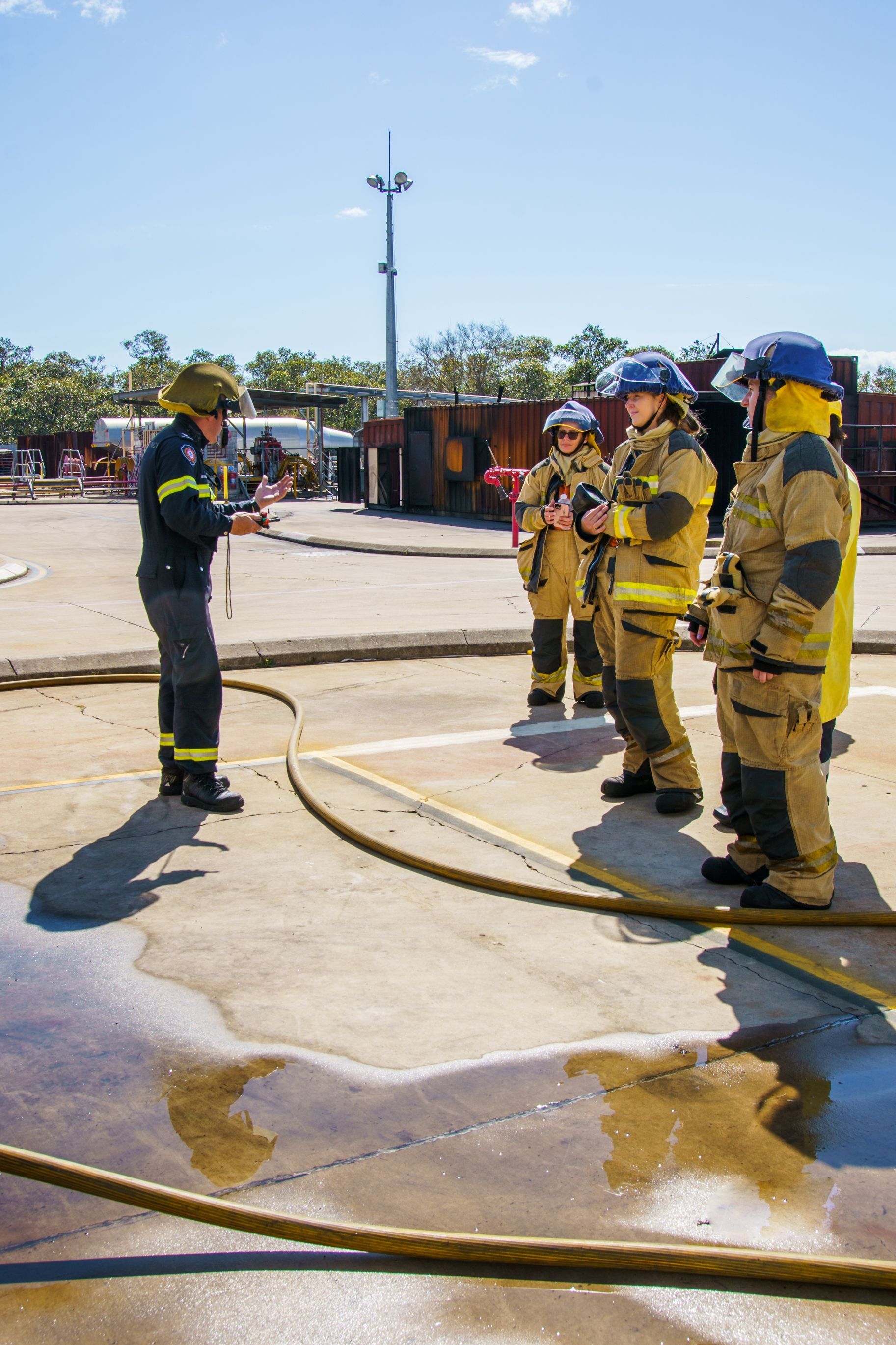 Firefighters in protective gear, listening to an instructor outdoors on a sunny day.
