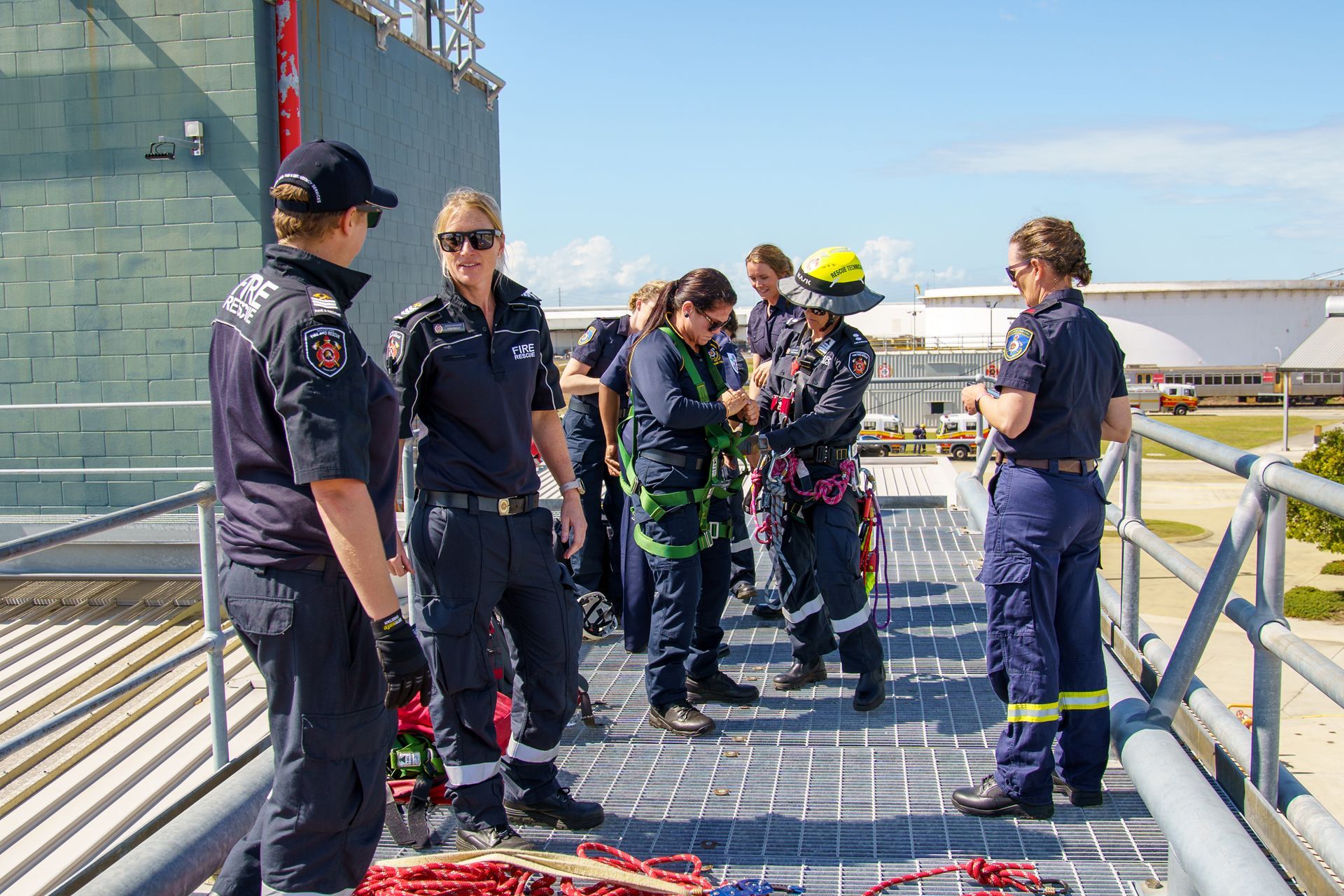 Emergency responders on a rooftop, one in harness. Sunny day. Blue uniforms. Discussing a rescue plan.