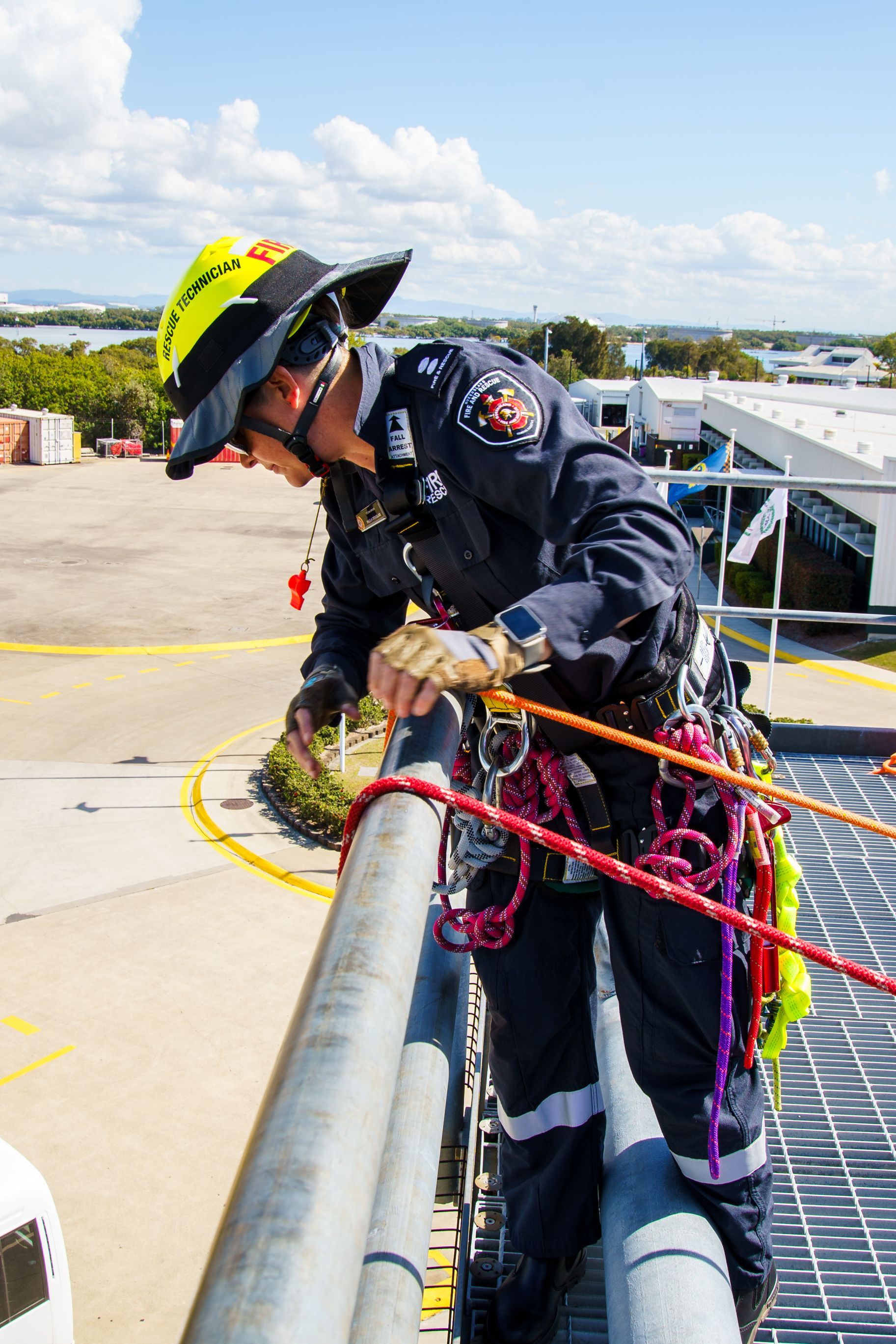 Firefighter in dark blue uniform, helmet, checking ropes on a rooftop with blue sky background.
