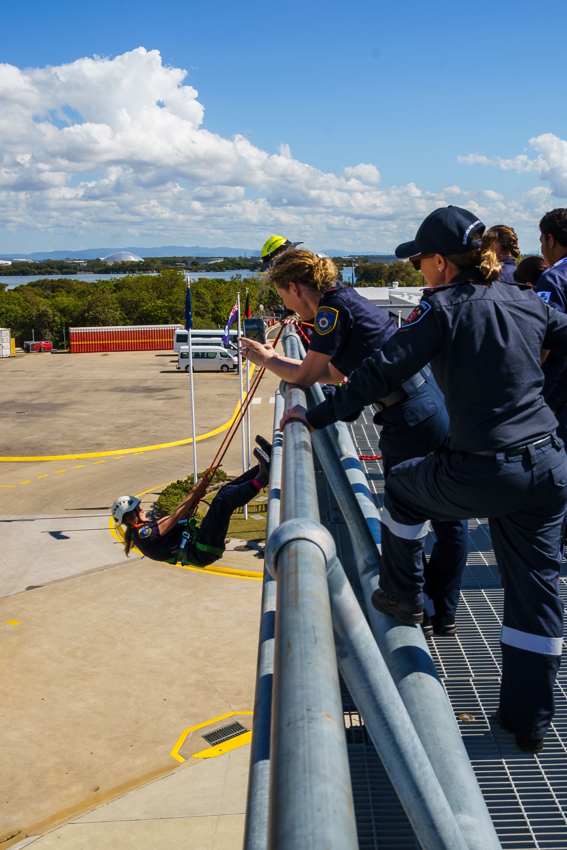 Firefighters training: one rappels down building, others watch from railing on sunny day.