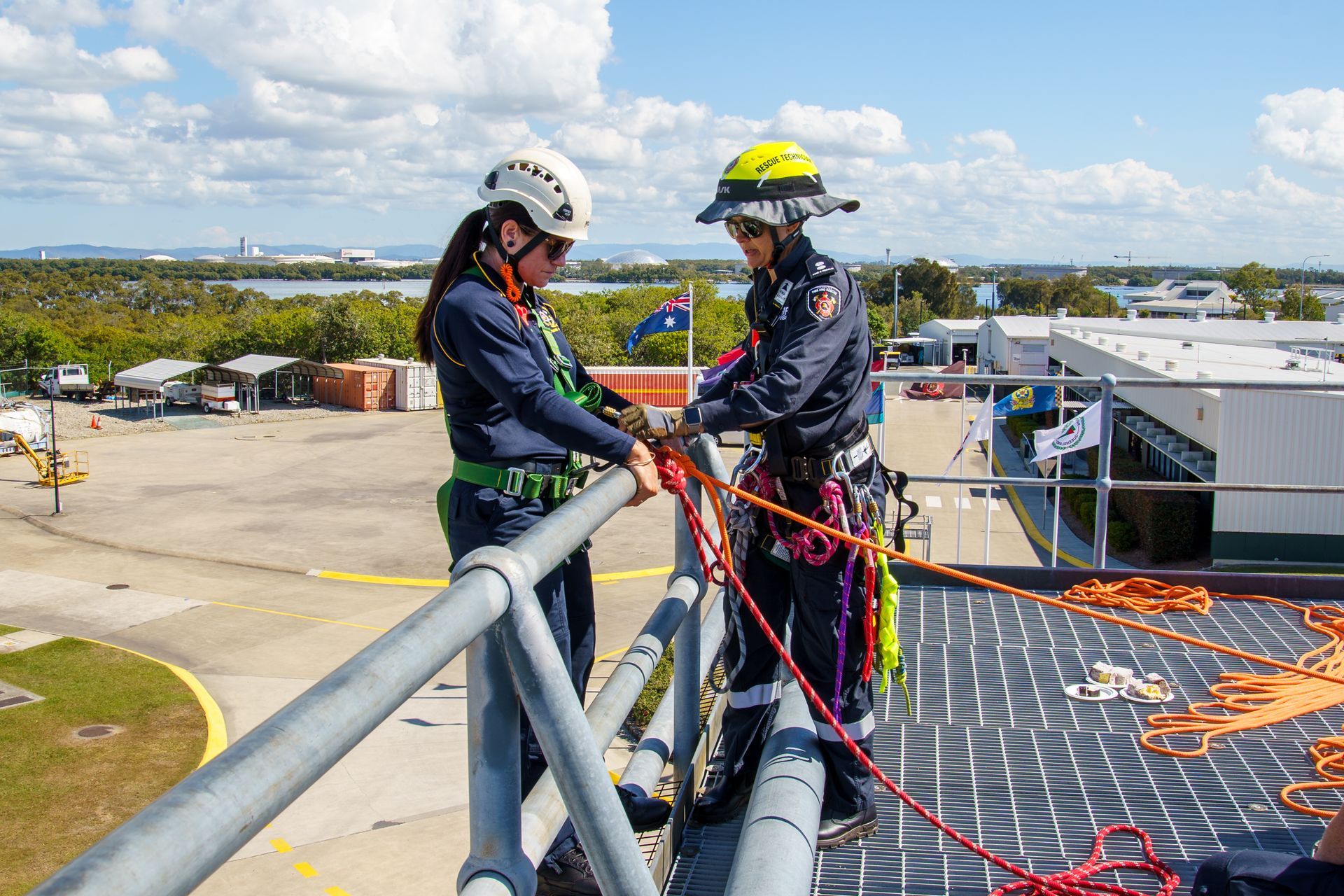 Two people in safety gear preparing ropes on a rooftop. Overcast sky, industrial setting.