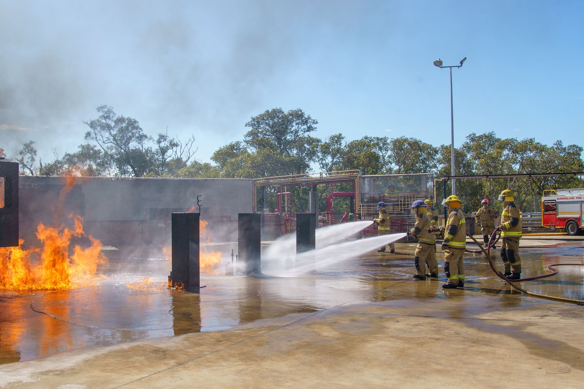 Firefighters training, using water hoses on a burning structure outdoors on a sunny day.